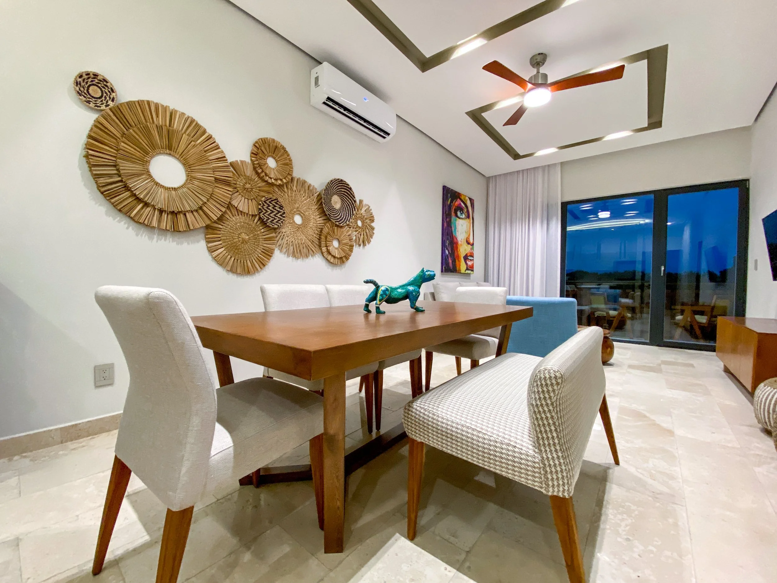 Una sala con mesa de madera, varias sillas blancas y decoraciones de mimbre en la pared, además de un cuadro colorido y una figura de dinosaurio sobre la mesa.