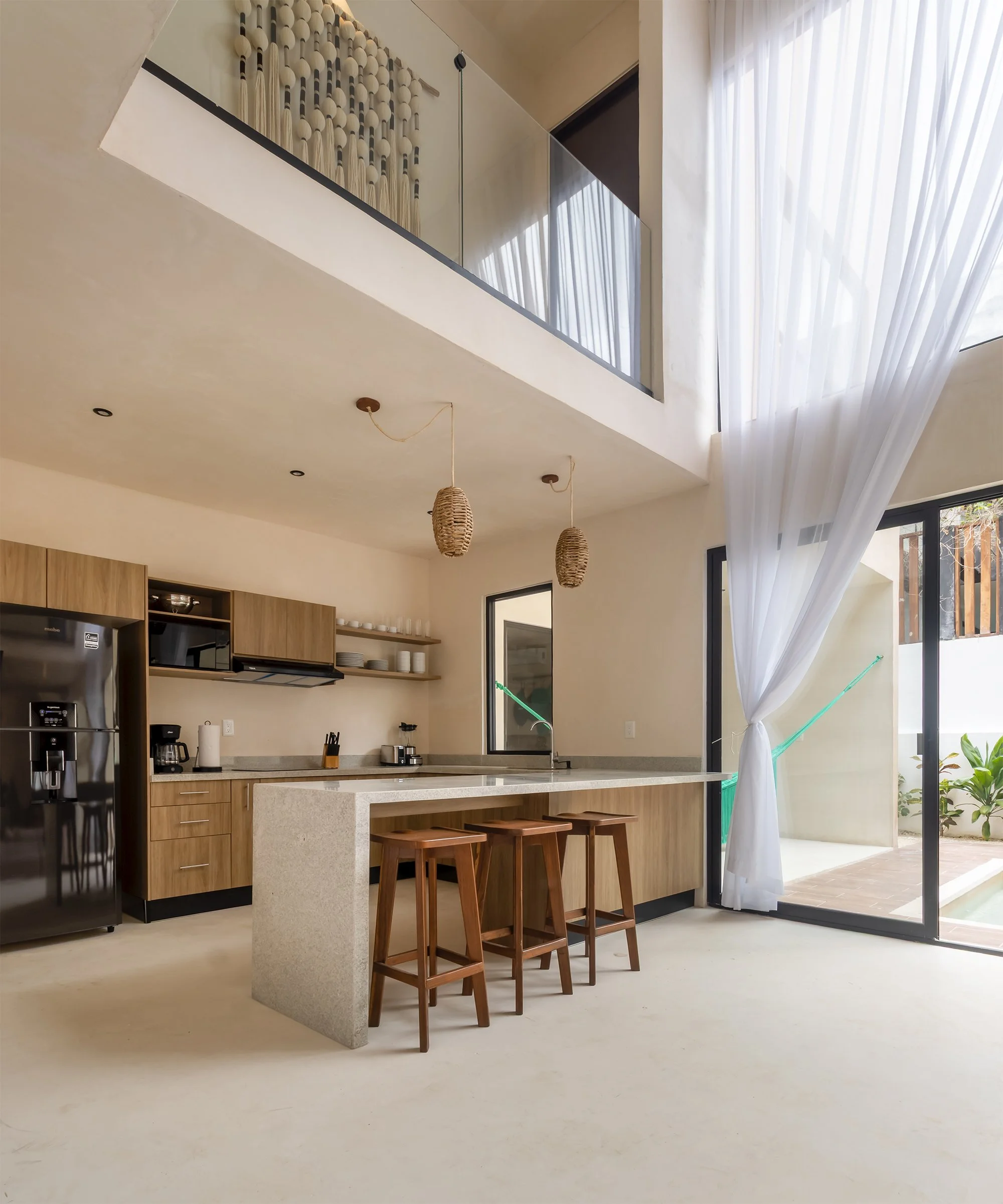 Cocina moderna con barra de granito, tres taburetes de madera, gabinetes de madera clara, nevera de acero inoxidable, estantería abierta con platos y tazas, paredes de color beige, cortinas blancas y grandes ventanas con vista al exterior.