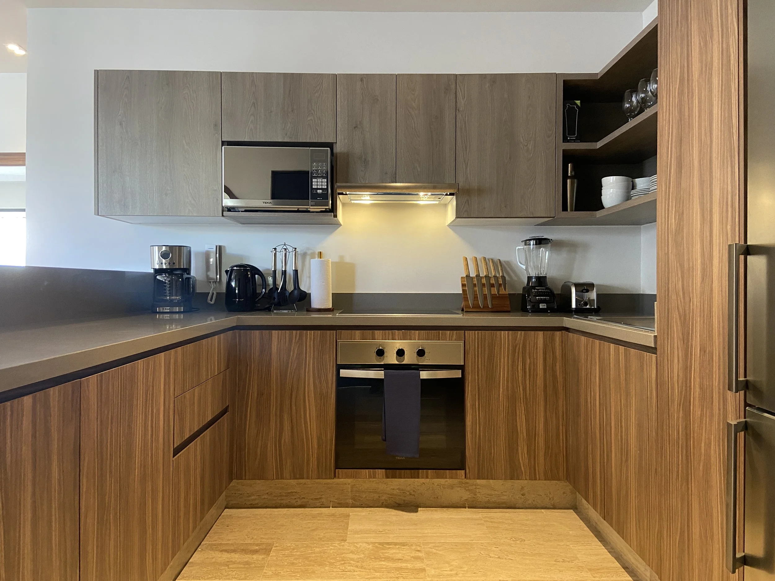 Cocina moderna con armarios de madera, microondas, cafetera, licuadora, utensilios y platos en estantes abiertos.