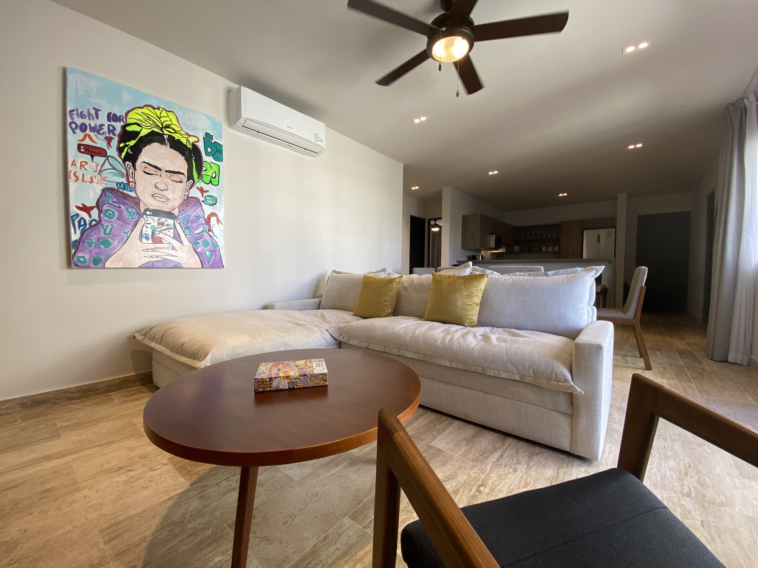 Sala de estar con un sillón grande de color beige y cojines amarillos, mesa redonda de madera con un rompecabezas y una silla de madera con asiento de malla negra. En la pared, un cuadro de un dibujo de una mujer con cabello con trenzas rubias, fondo azul y texto que dice "Fight for Power," entre otros. Hay un ventilador de techo y un aire acondicionado en la pared blanca.