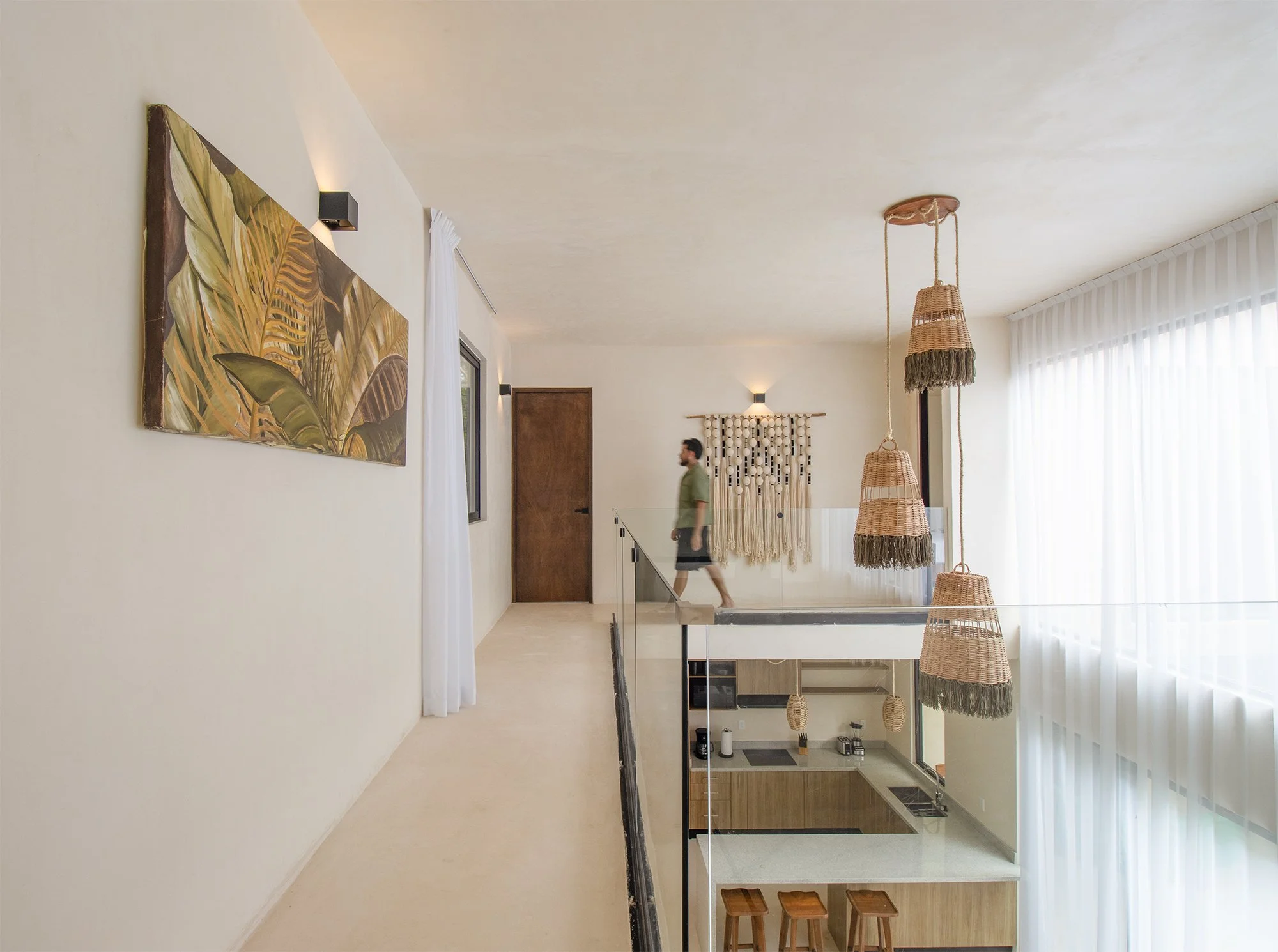 Interior de una casa moderna con paredes blancas, cortinas blancas, lámparas de mimbre colgantes, y una pared decorativa con bolas blancas. Una persona caminando por la sala y una cocina visible debajo de una escalera de vidrio con asientos de madera.