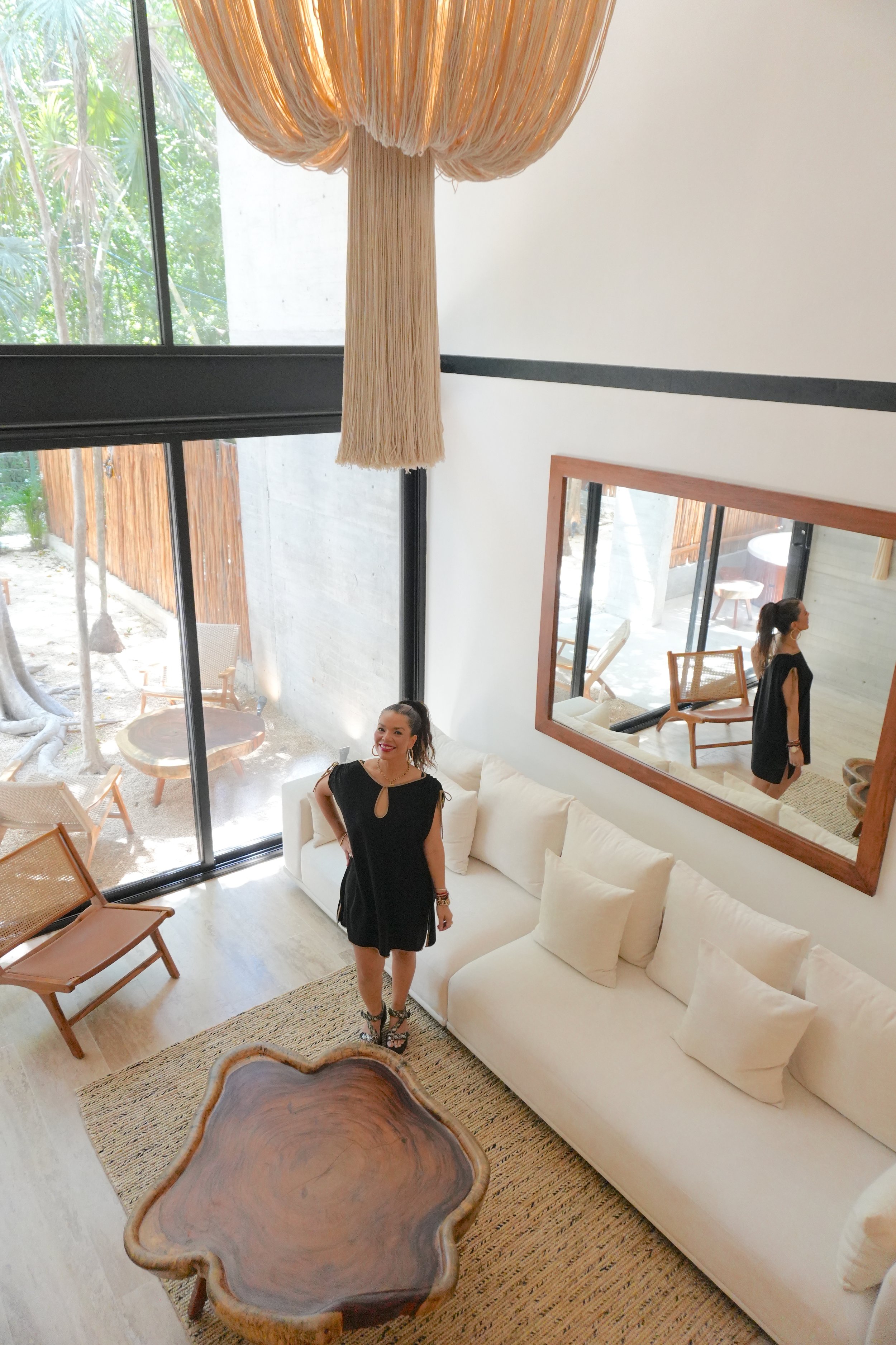 Interior de una sala moderna con sillones blancos, mesa de madera en forma irregular, mujer con vestido negro posando, espejo grande en la pared, ventanas grandes con vista a un jardín, cortinas de seda, muebles de madera, ambiente luminoso y acogedor.