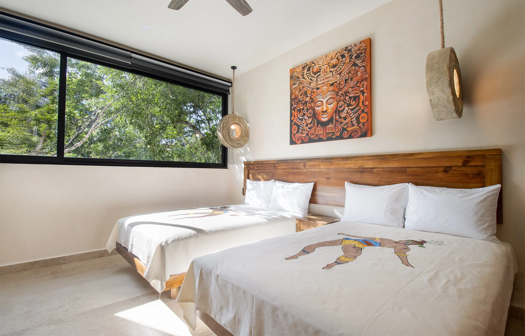 Dos camas dobles con sábanas blancas, una con una manta con dibujo de un tico y la otra con un arte en la pared que representa una figura indígena. Hay una ventana grande con vista a árboles verdes, una lámpara de pared de piedra y un ventilador en la habitación.