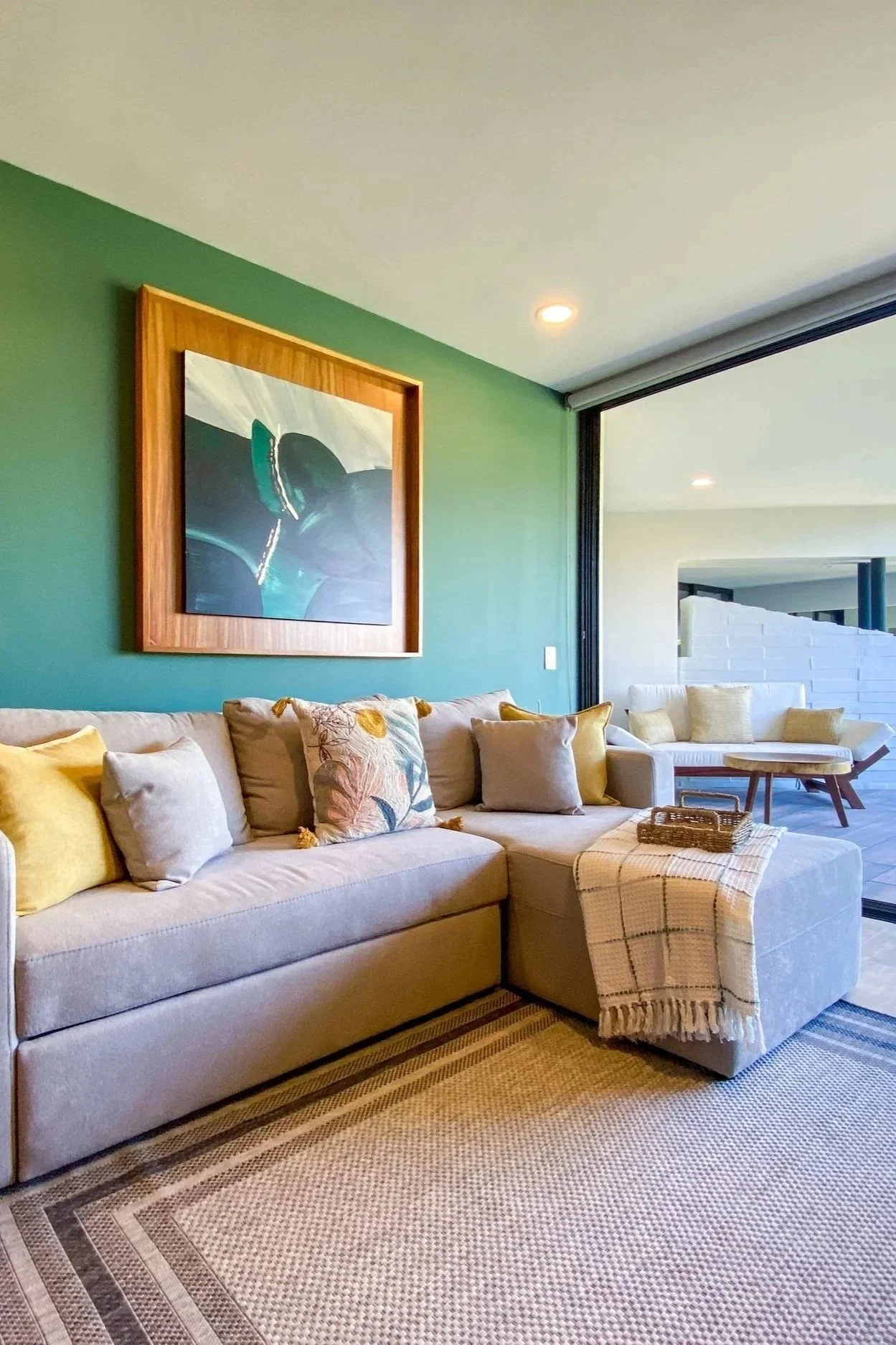 Sala de estar con sillón beige, cojines decorativos en tonos neutros y amarillos, alfombra de colores tierra, pared verde con cuadro abstracto y amplio ventanal con cortinas blancas.
