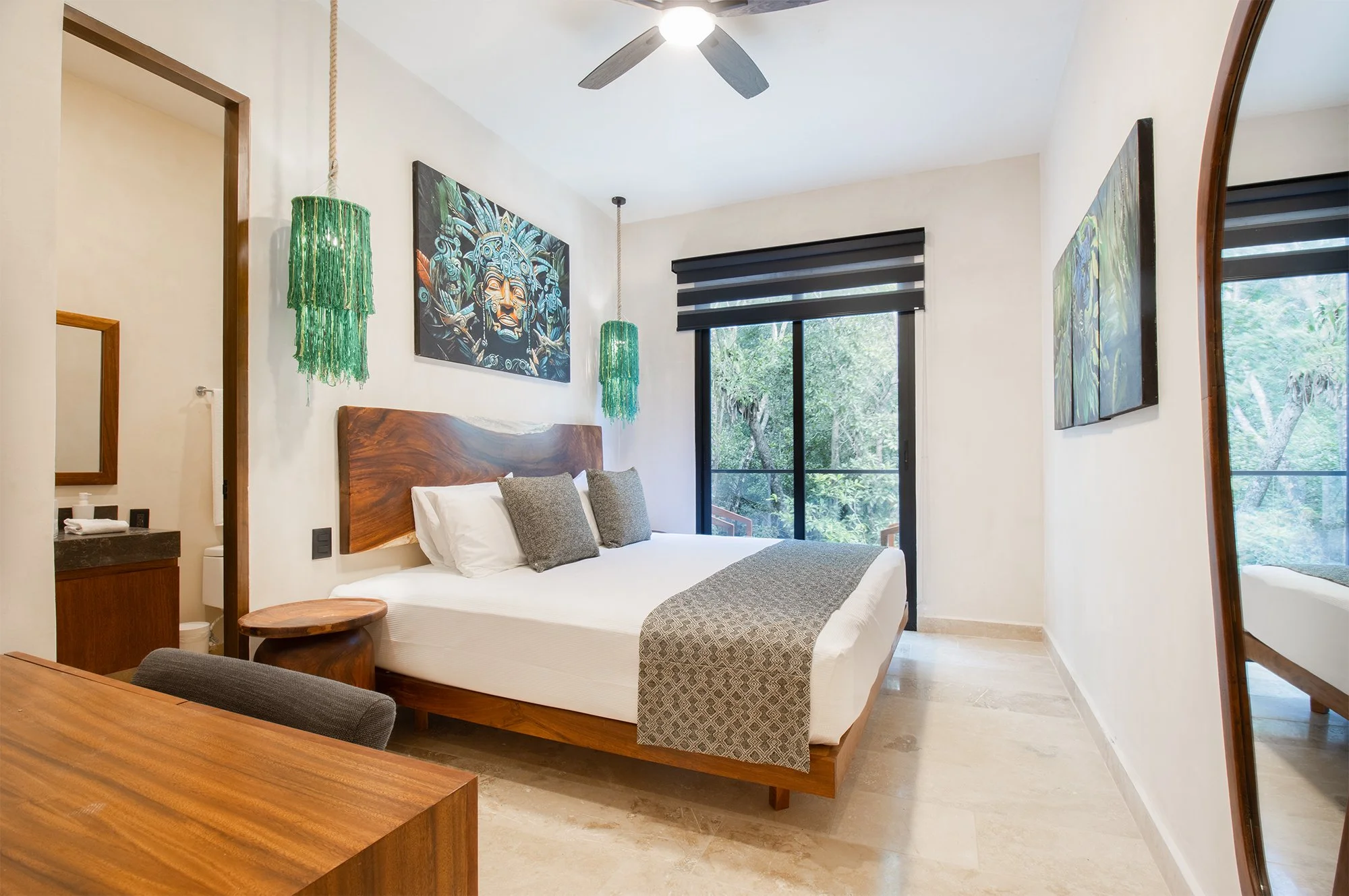 Habitación moderna con cama de madera, arte mural con rostro mayacánico, ventanas con vista a vegetación, ventilador en el techo y decoración minimalista.