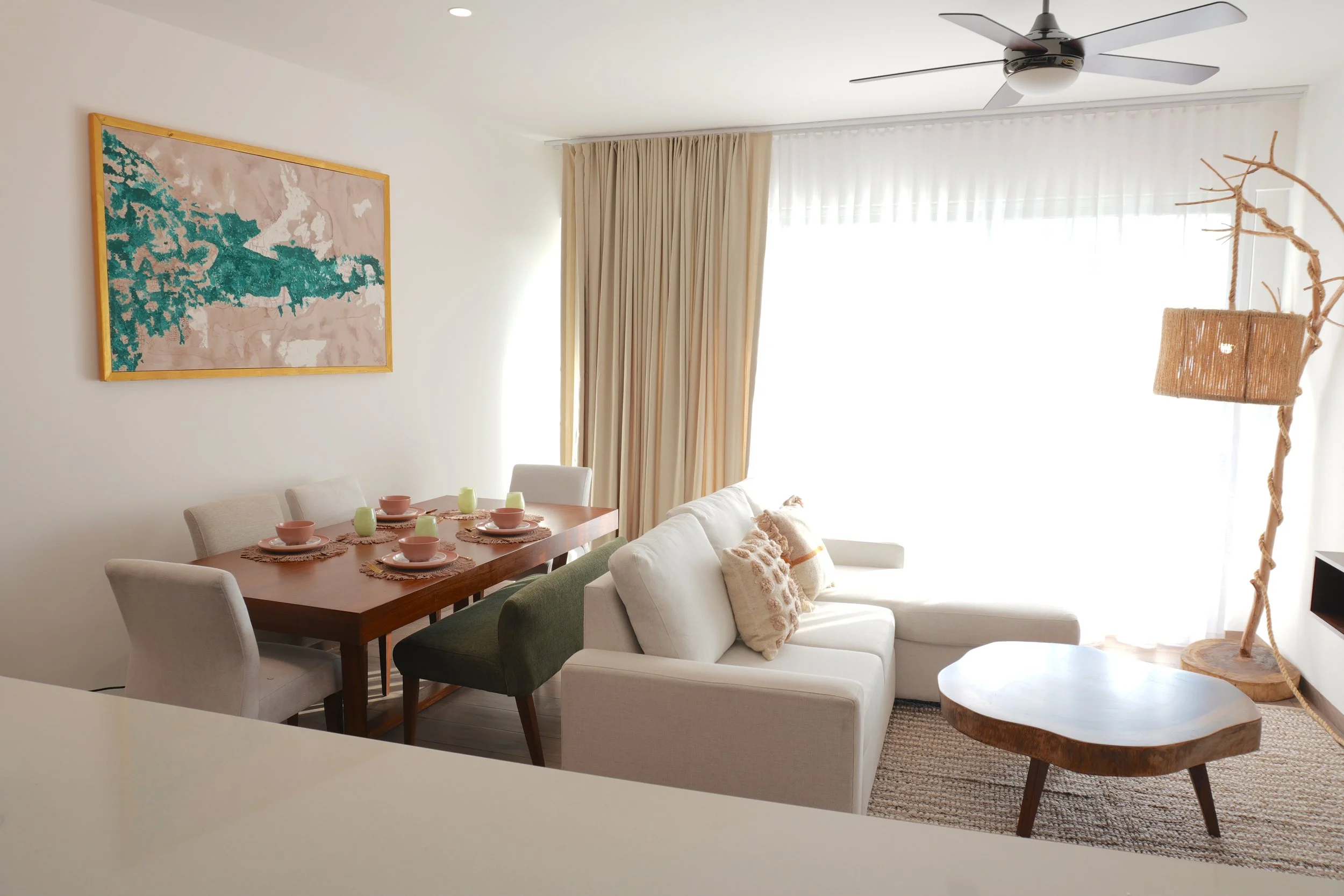 Sala de estar con mesa de comedor, sillones blancos y verdes, cortinas beige, un ventilador de techo, una lámpara de mimbre y una pintura abstracta en la pared.