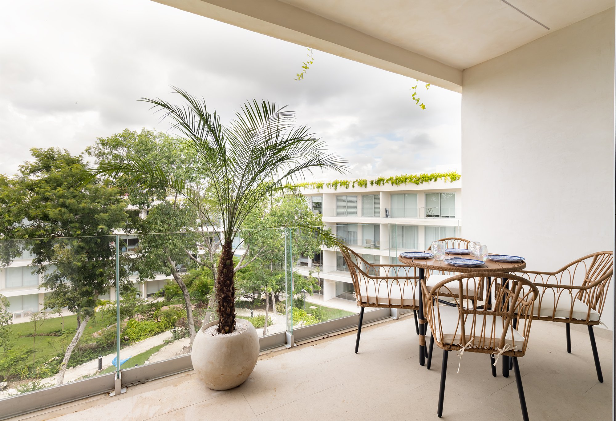 Balcón con mesa y sillas de mimbre, planta en maceta grande, árboles y edificios de apartamentos en el fondo, cielo nublado.
