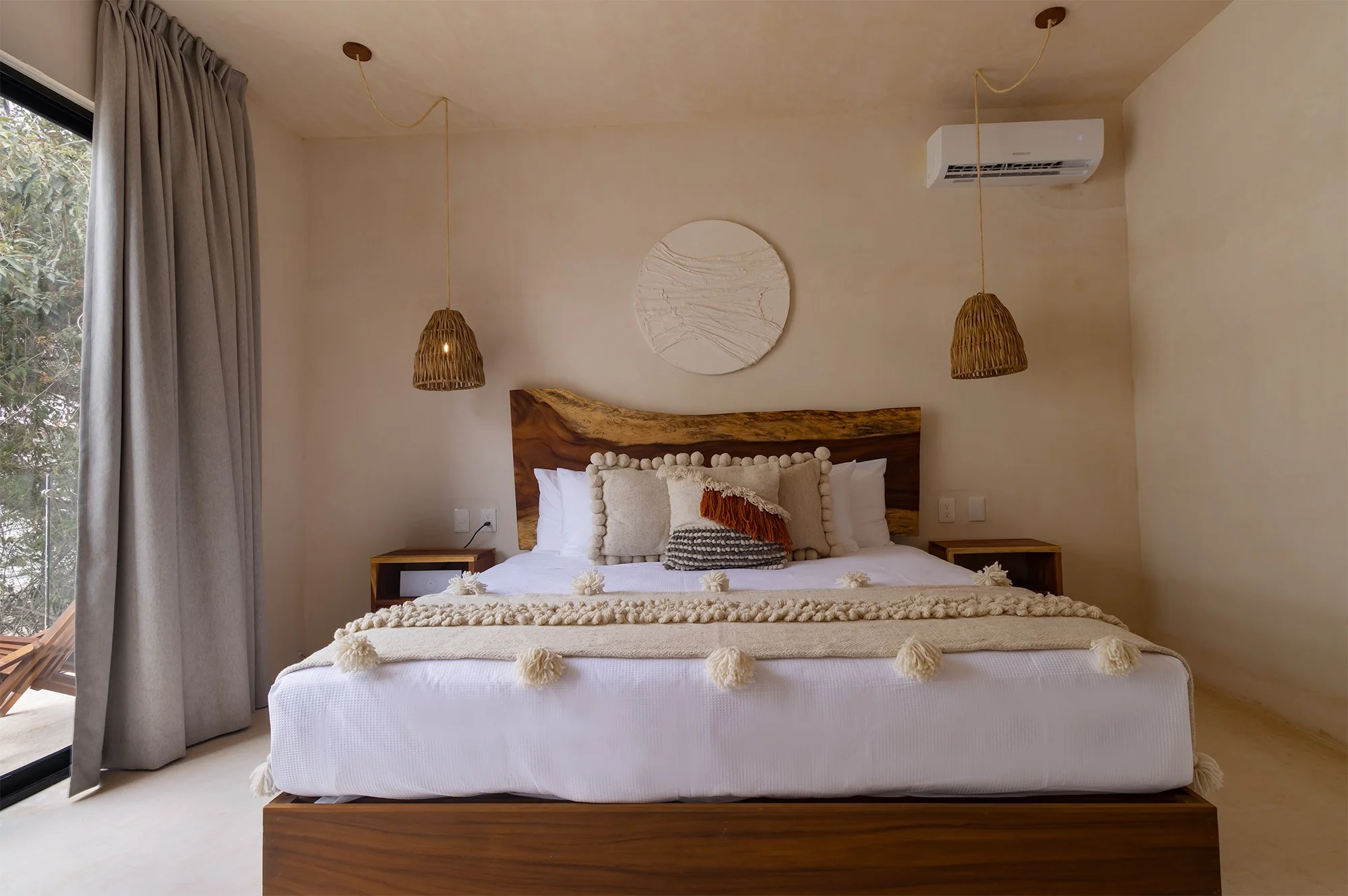Dormitorio con cama grande, cabecera de madera, cojines decorativos y cortinas grises junto a una ventana de vidrio cercano a plantas exteriores, con lámparas de sujeción de cuerda en el techo.
