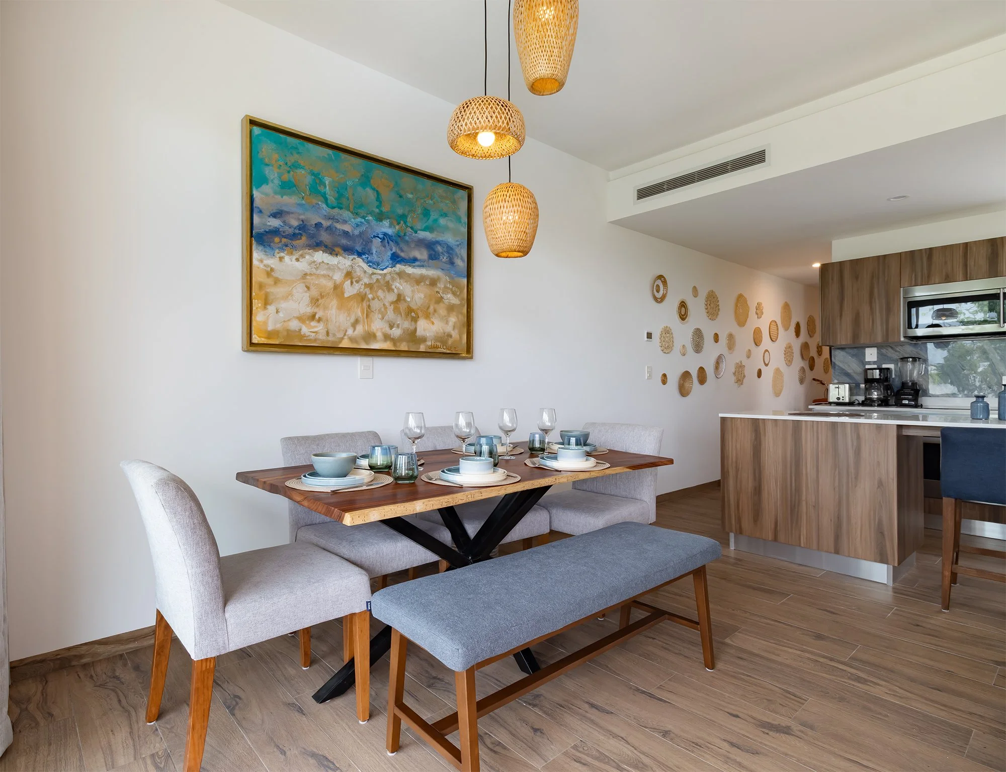 Comedor con mesa de madera, sillas tapizadas en tonos gris y una banca de madera con cojines, decoraciones en la pared con cuadros y objetos decorativos de madera, cocina moderna con gabinetes de madera, electrodomésticos y ventanas grandes, lámparas de mimbre colgantes.
