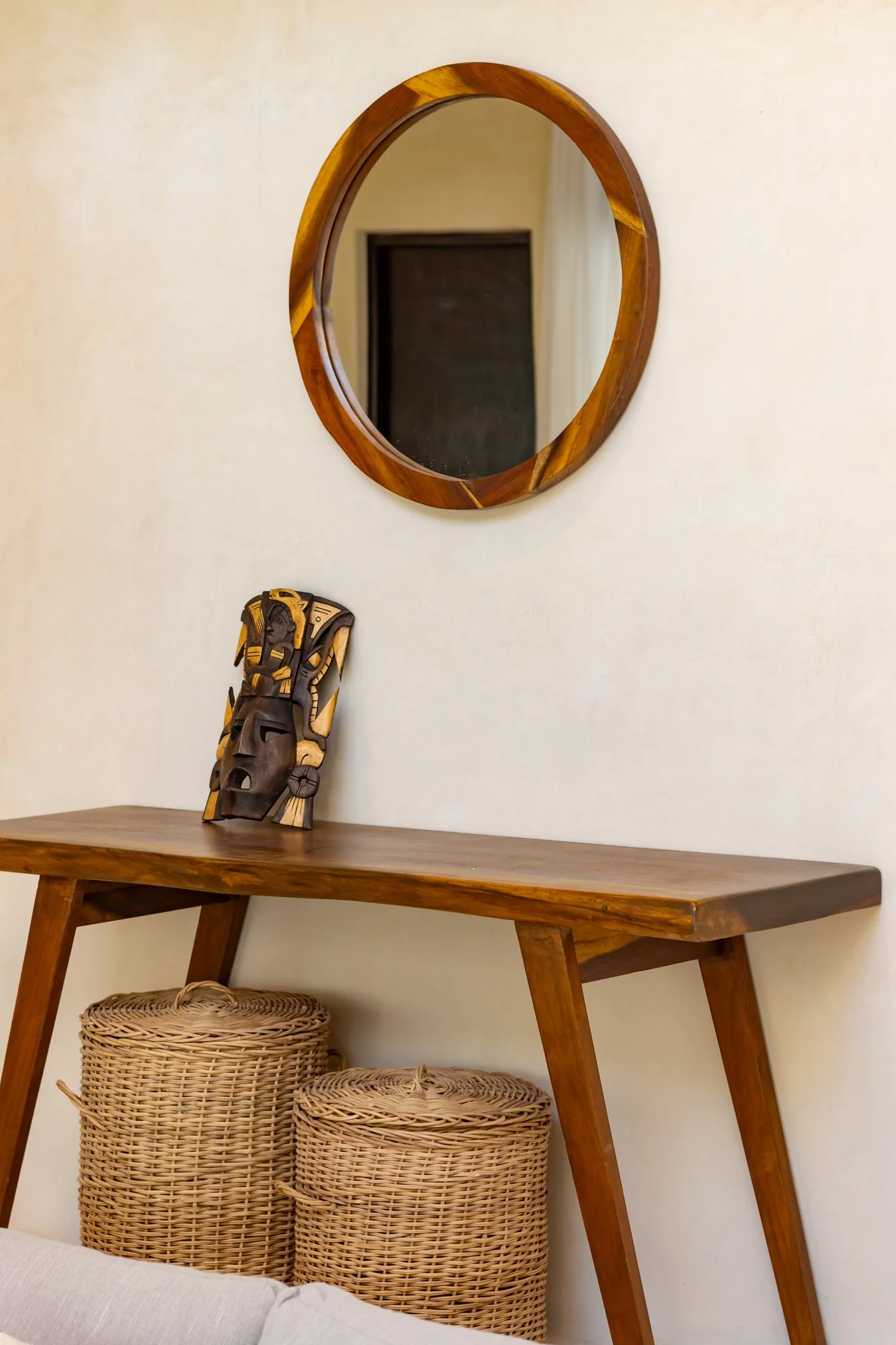 Espejo redondo de madera en la pared, mesa de madera con una máscara africana, cestas de mimbre debajo de la mesa.