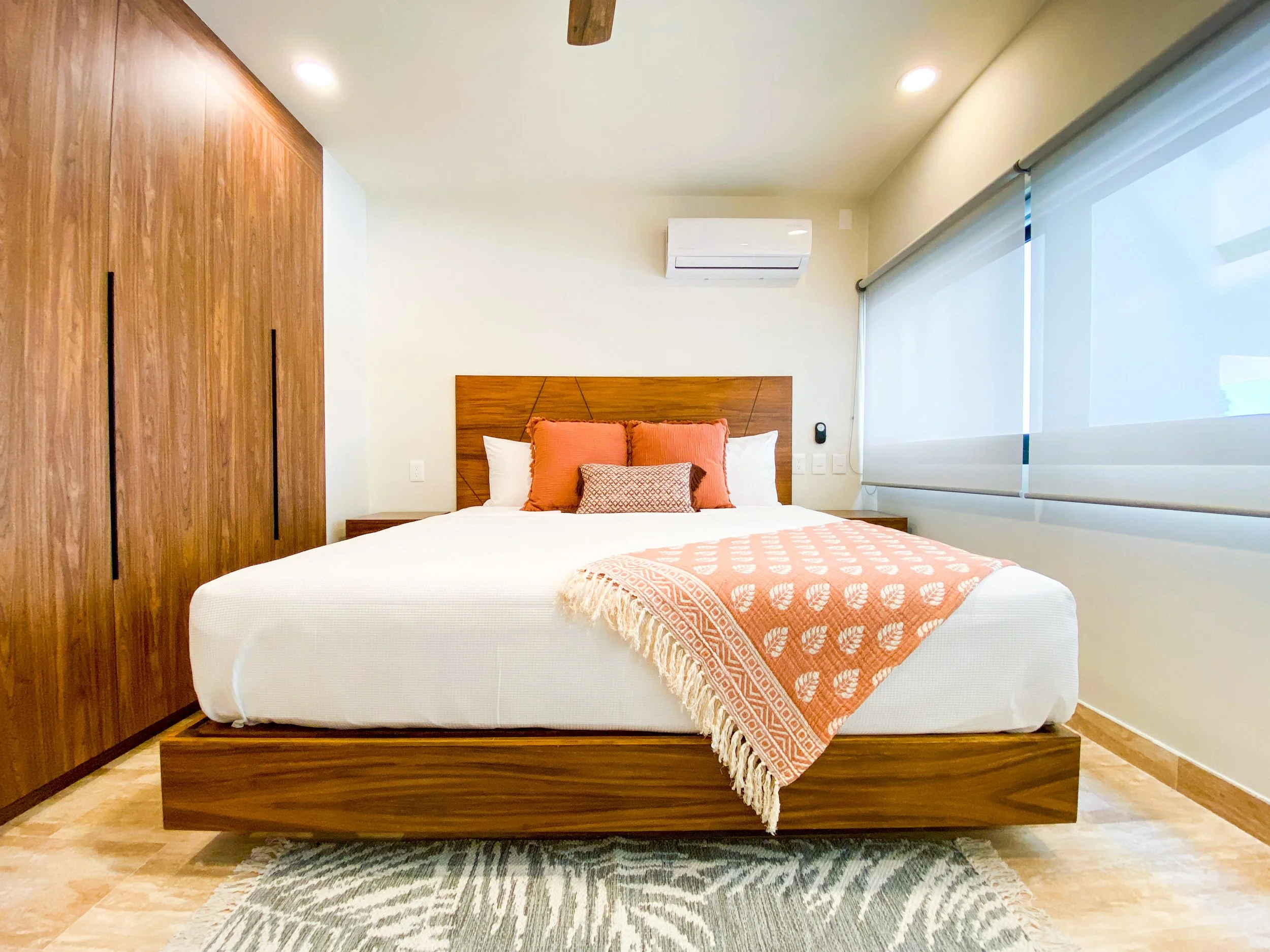 Habitación moderna con cama grande, cabecera de madera y accesorios en tonos naranja, suelo de madera y cortinas blancas enrollables.