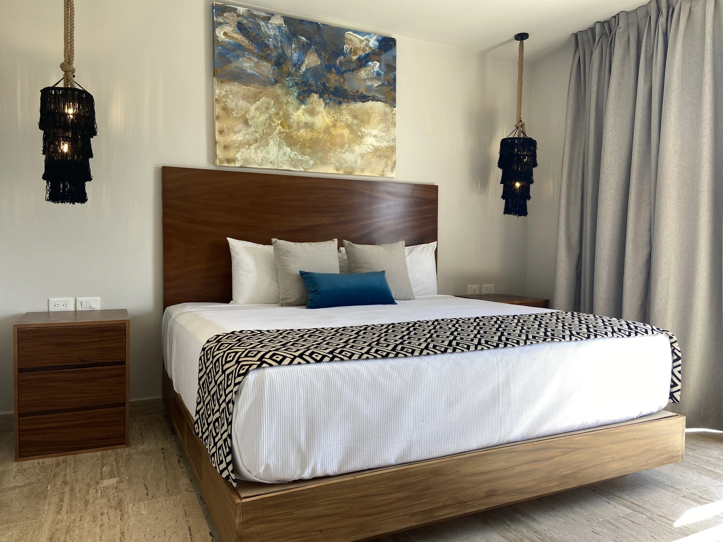 Habitación moderna con cama grande, ropa de cama blanca con detalles en negro, almohadas blancas y una almohada azul. Cuatro mesas de noche de madera, lámparas colgantes negras y un cuadro abstracto en la pared. Cortinas gris claro cubren la ventana.