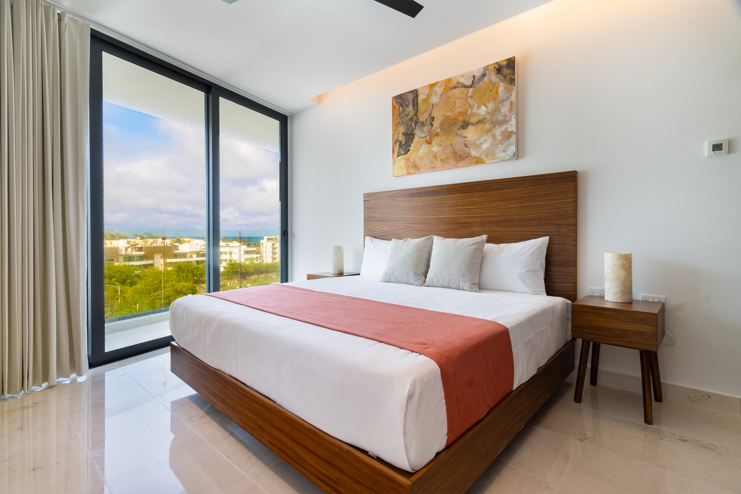 Habitación con cama moderna de madera, almohadas blancas, edredón rosa, vista por ventana grande con cortinas blancas hacia edificios y naturaleza.