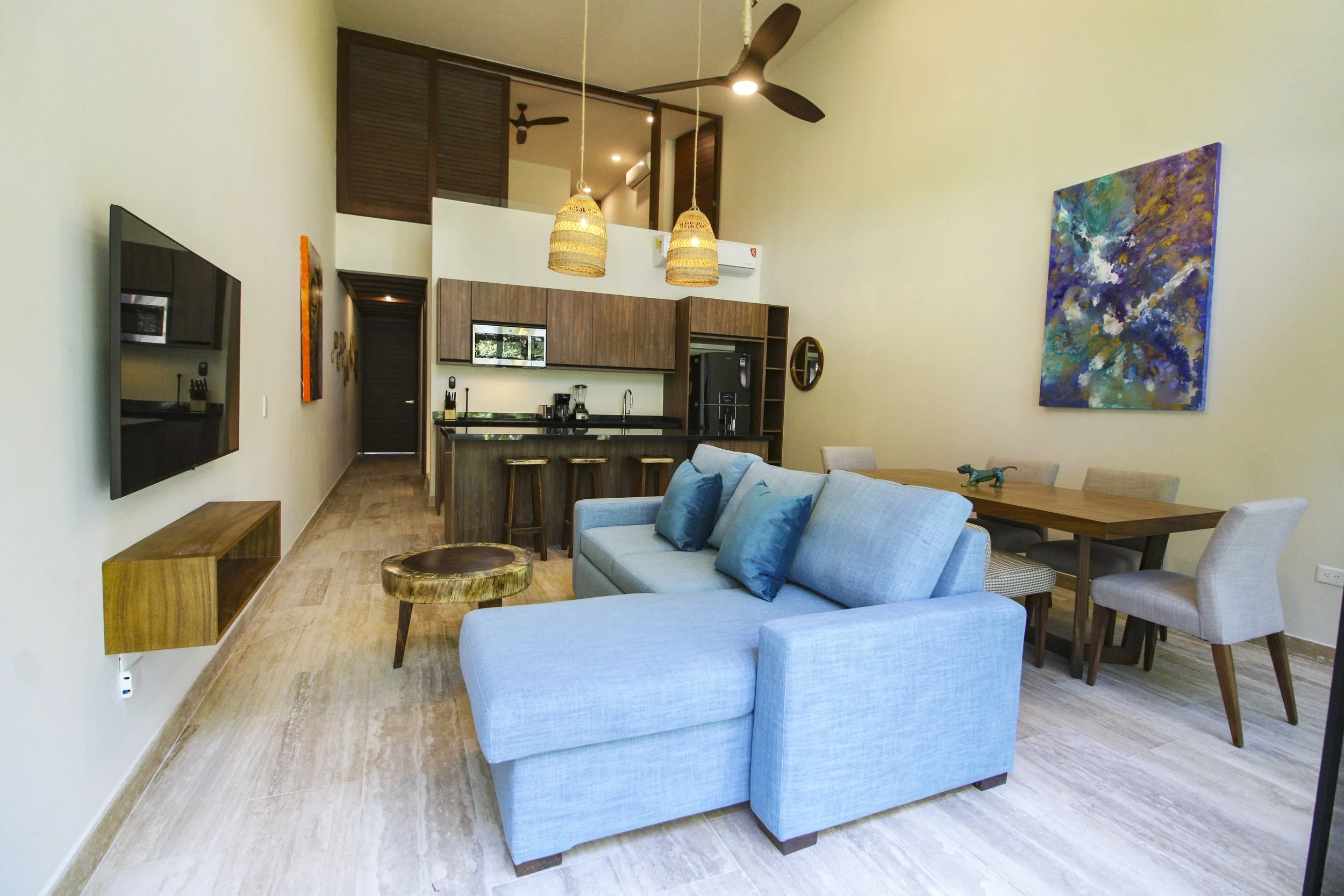 Sala de estar moderna con sofá azul, comedor con sillas de madera, una mesa y arte abstracto en la pared. Cocina en el fondo con gabinetes de madera, isla con taburetes y electrodomésticos, ventiladores y lámparas colgantes de mimbre.