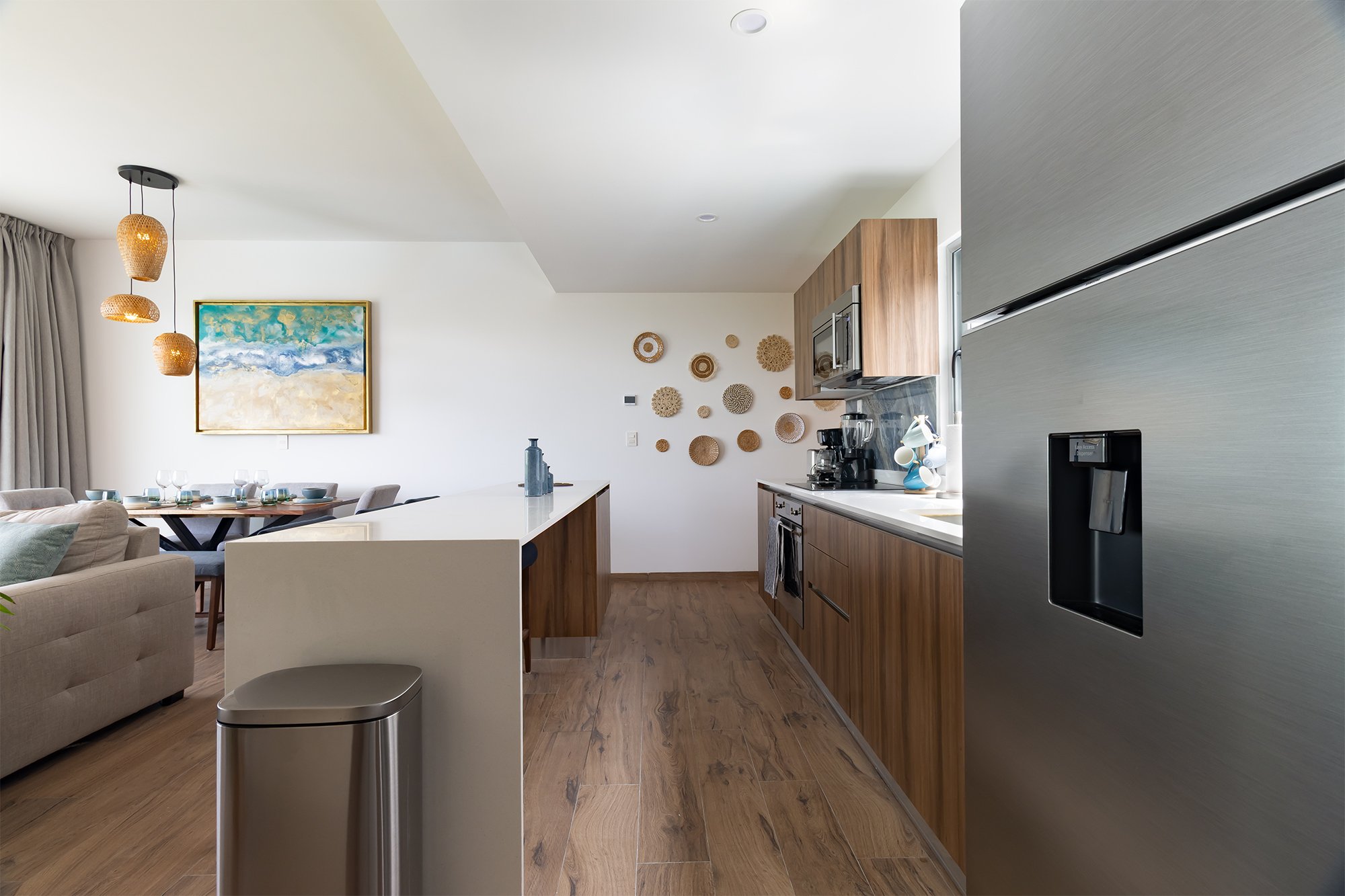 Cocina moderna con armarios de madera, electrodomésticos de acero inoxidable, paredes blancas decoradas con cestas de mimbre, y una pared con arte de cuadros de color azul y beige.