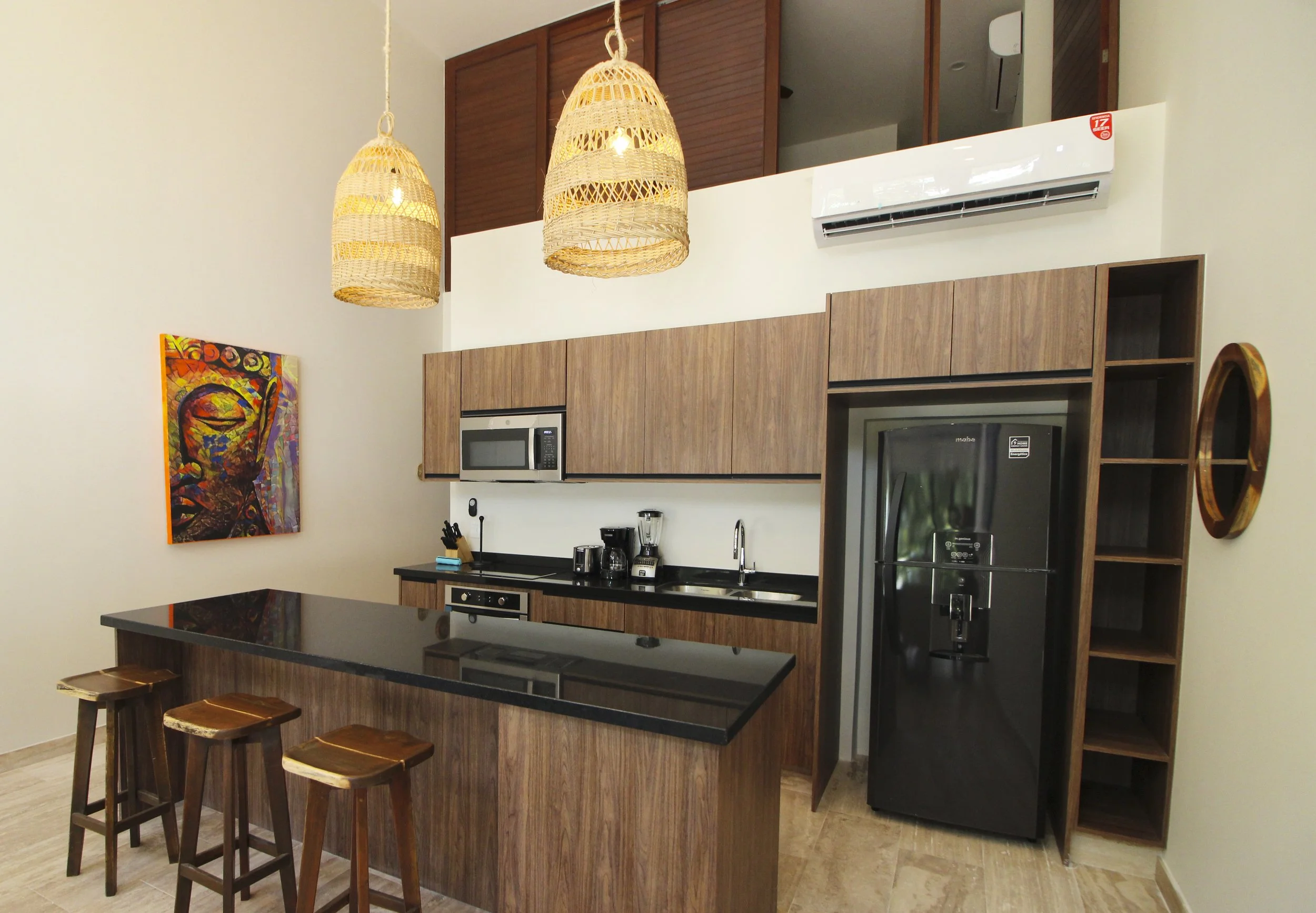 Cocina moderna con paredes blancas, gabinetes de madera, una nevera negra, microondas integrado, electrodomésticos de cocina, una encimera oscura, dos taburetes de madera y lámparas colgantes de mimbre.