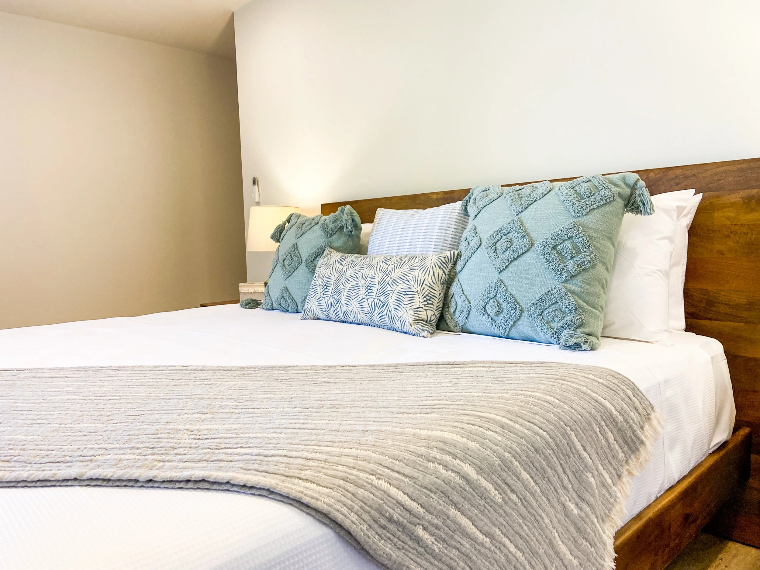 Cama con almohadas decorativas en tonos azul y beige, lámpara de mesa a la izquierda, pared blanca de fondo.