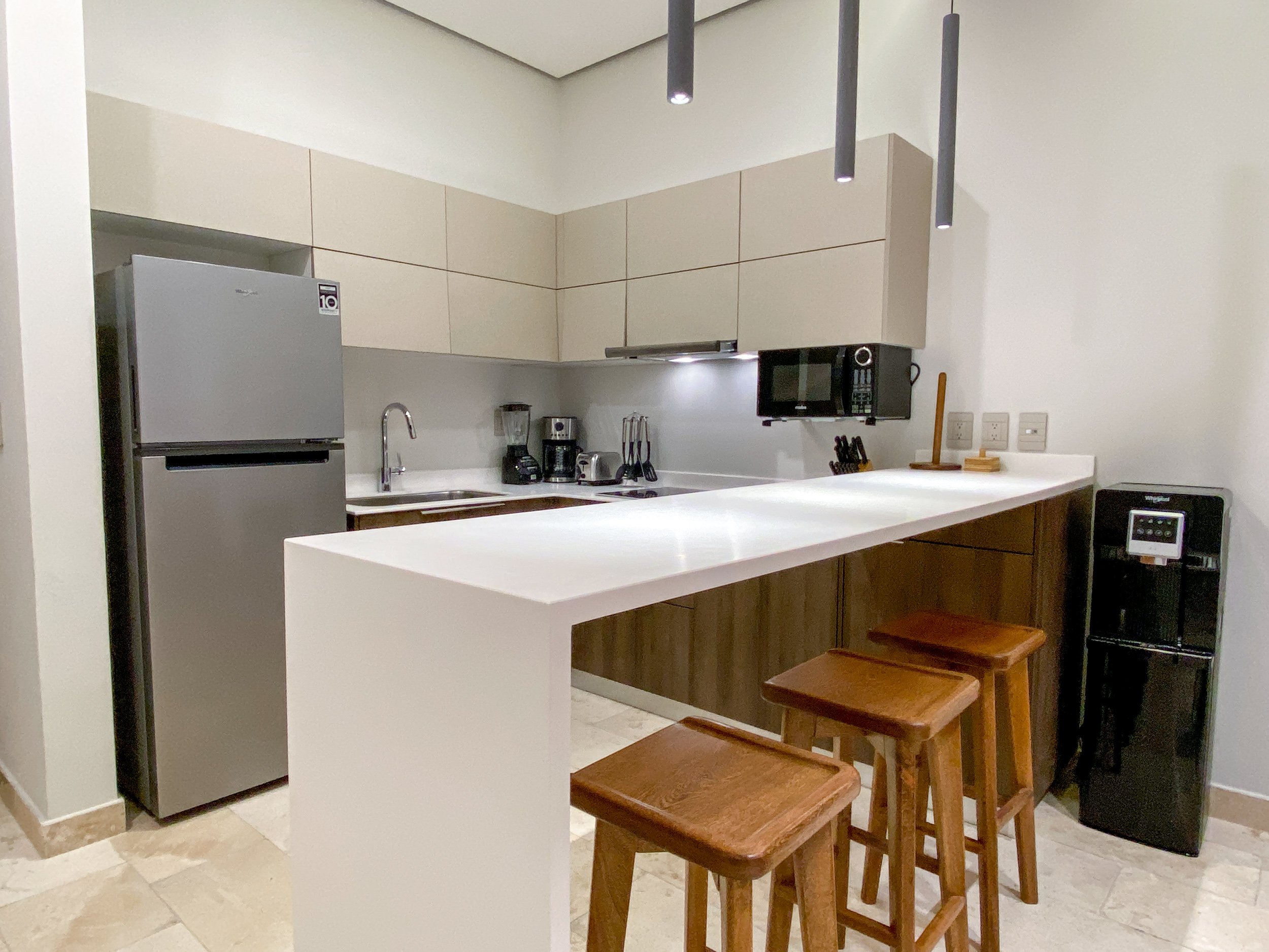 Cocina moderna con electrodomésticos en acero inoxidable, encimera de piedra, y área de barra con bancos de madera.