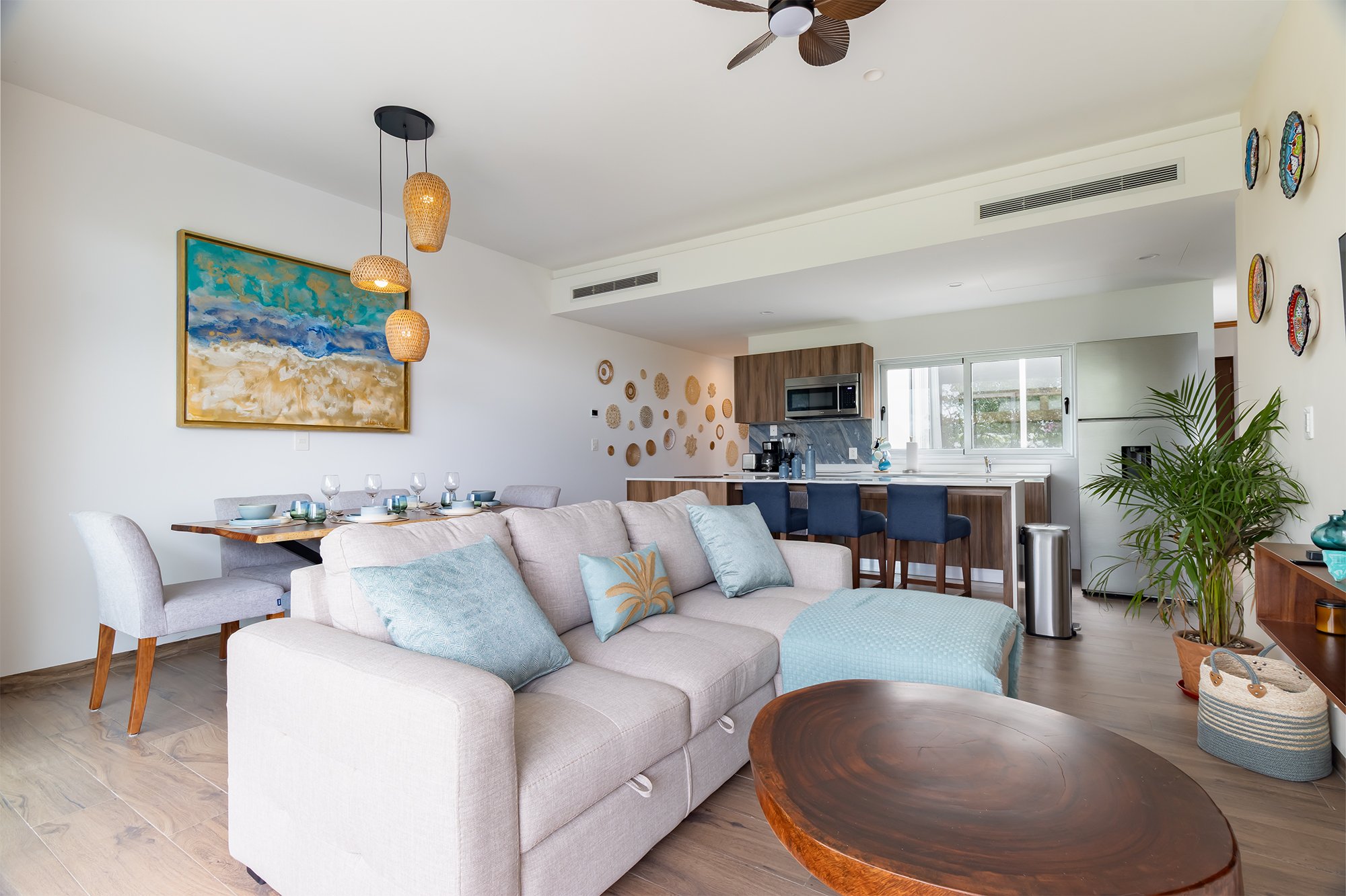 Sala de estar con sofá blanco, almohadas color pastel, mesa de centro de madera, comedor con sillas grises y casa con cocina moderna en el fondo, decorada con cuadros, plantas y lámparas de diseño.