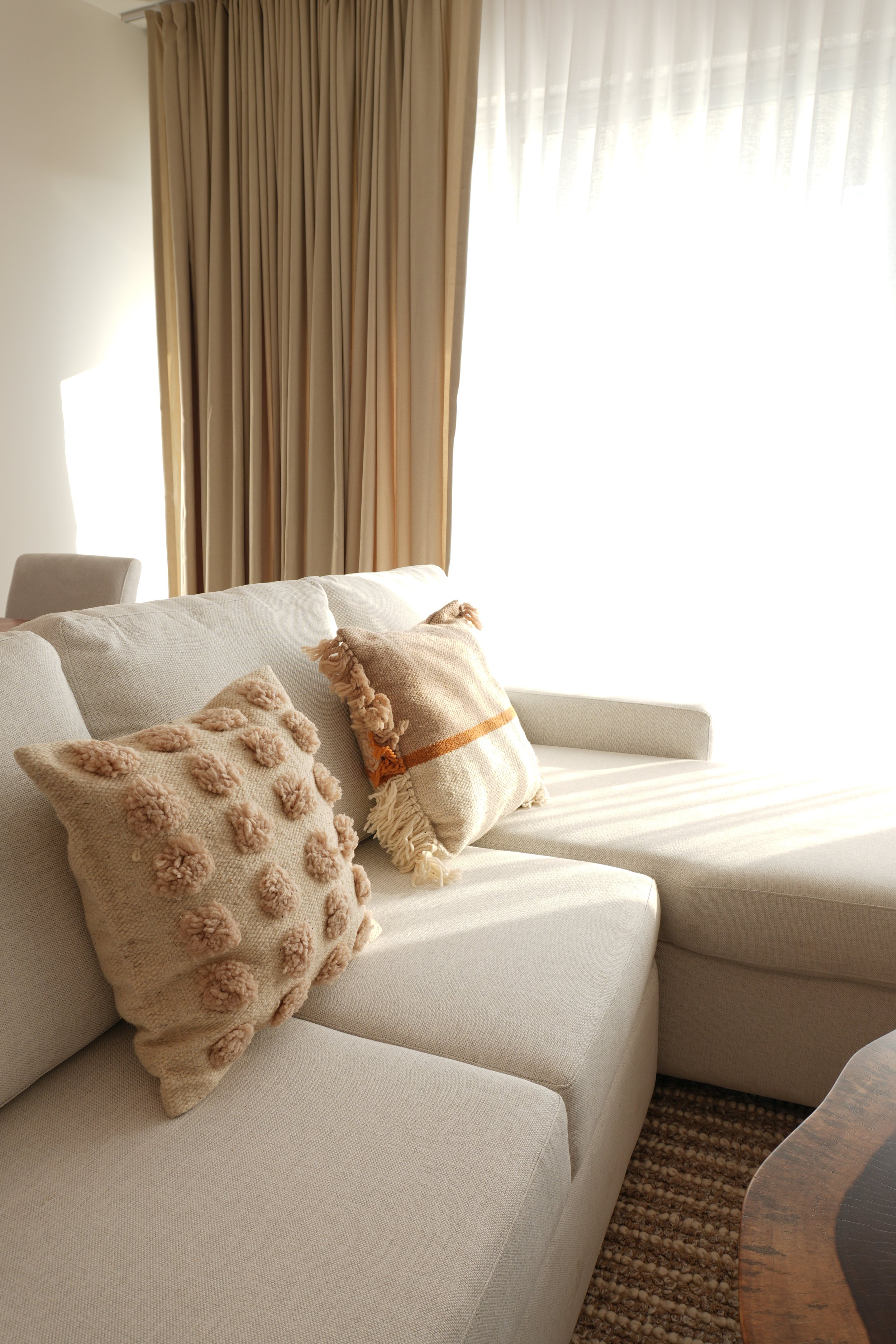 Sala de estar con sofá beige decorado con cojines de diferentes texturas y colores, cortinas beige y cortinas traslúcidas que dejan entrar luz natural, y una mesa de madera con borde recto y acabado oscuro, sobre una alfombra de diseño anudado en tonos marrones.