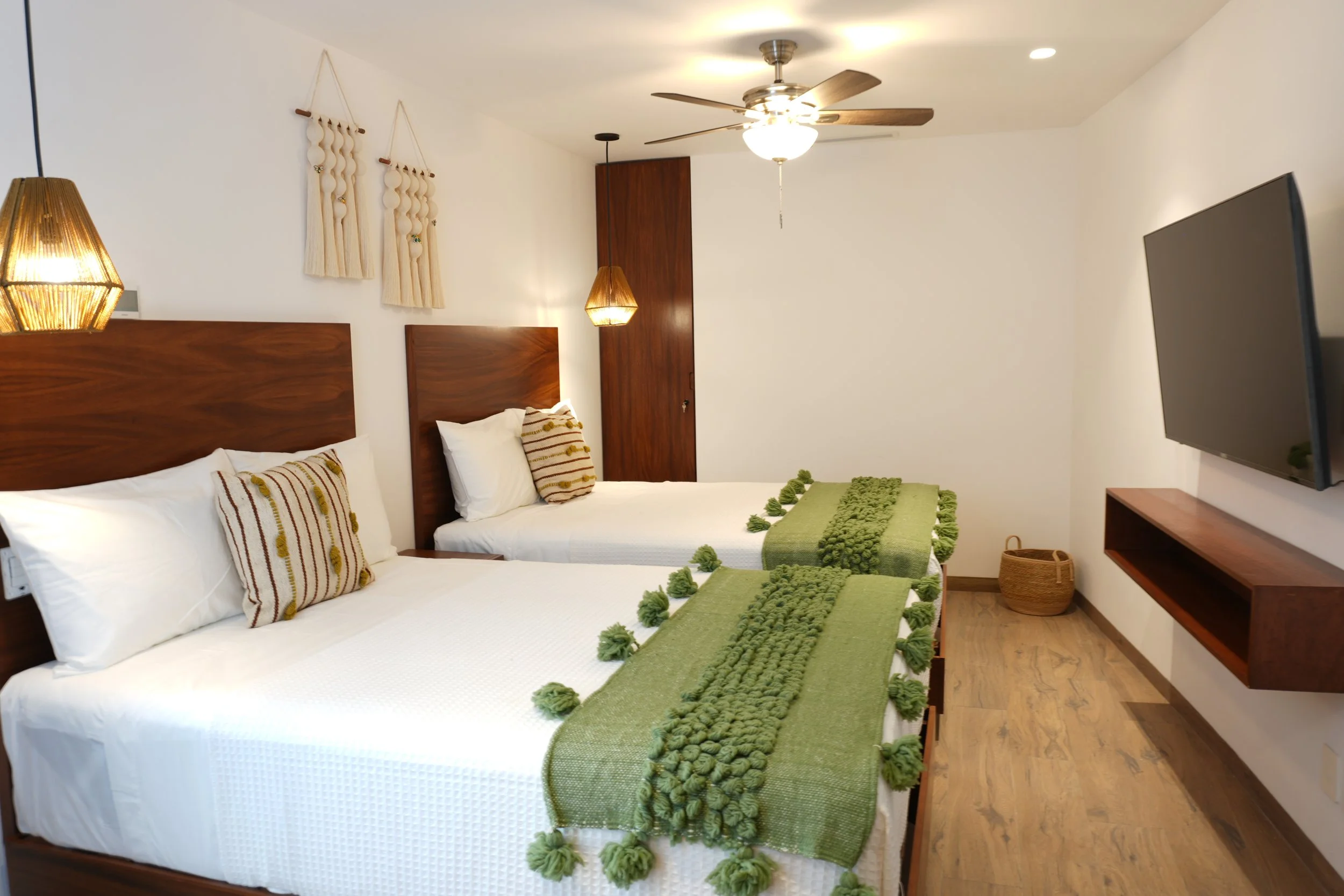 Dos camas con cabeceras de madera, mantas verdes decoradas con pompoms, almohadas con rayas. Iluminación cálida, paredes blancas, decoración de tela en la pared, ventilador en el techo, televisor en la esquina, suelo de madera.