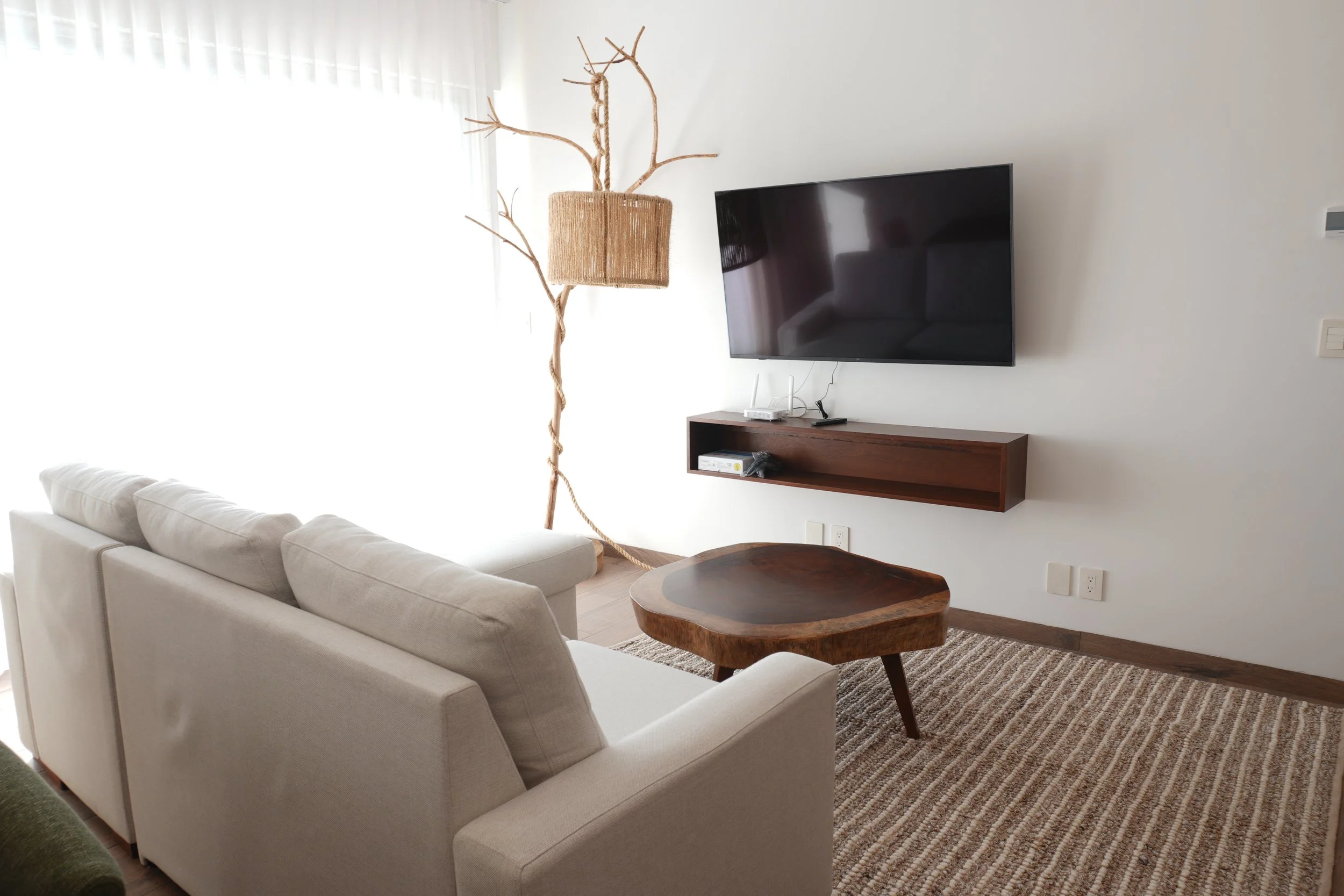 Sala de estar con sillones blancos, una mesa de madera, televisor de pantalla plana en la pared, lámpara de decoración en forma de árbol y cortinas blancas.