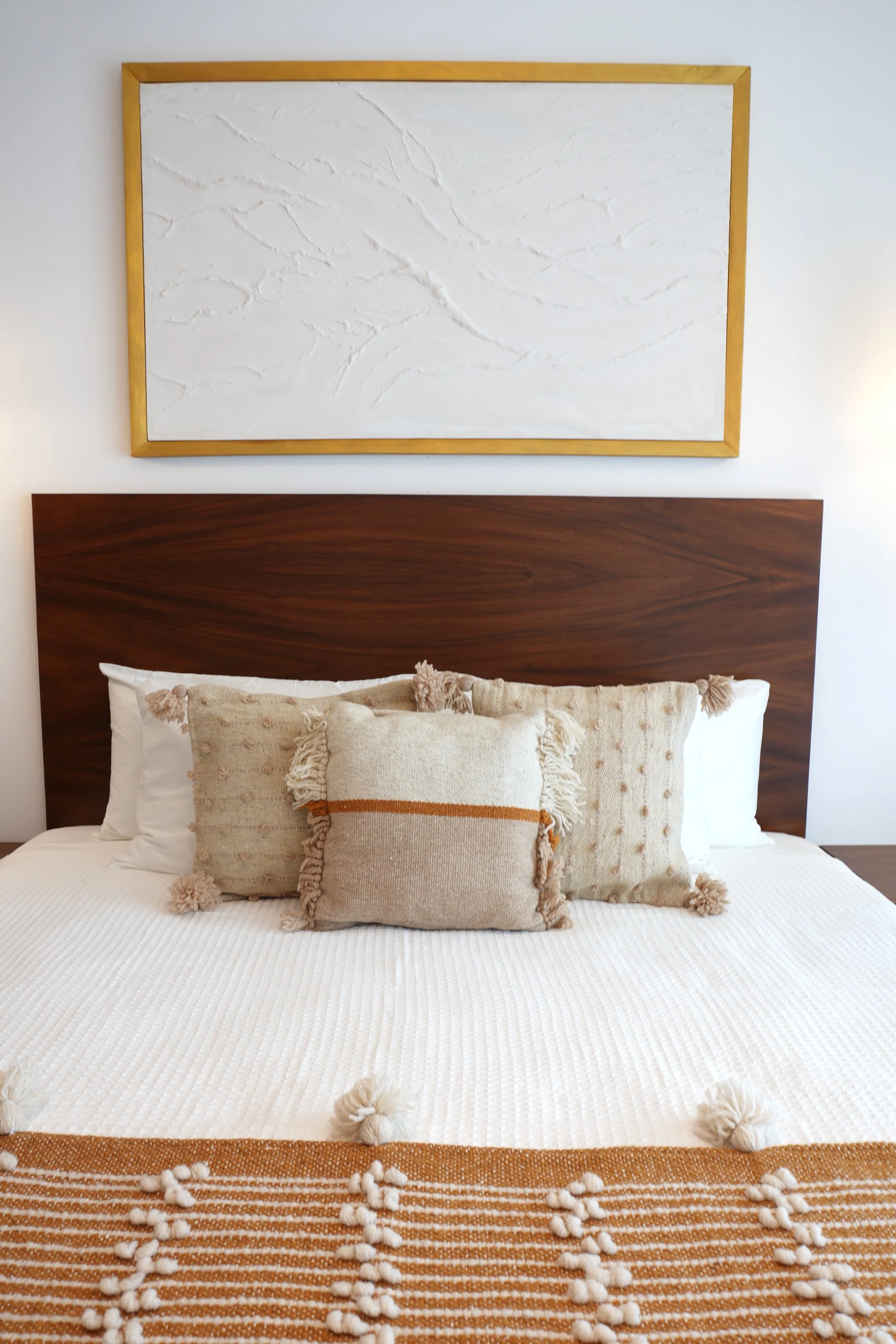 Cama con almohadas decorativas en tonos beige y textura tejida, madera oscura del cabecero y cuadro con textura en la pared
