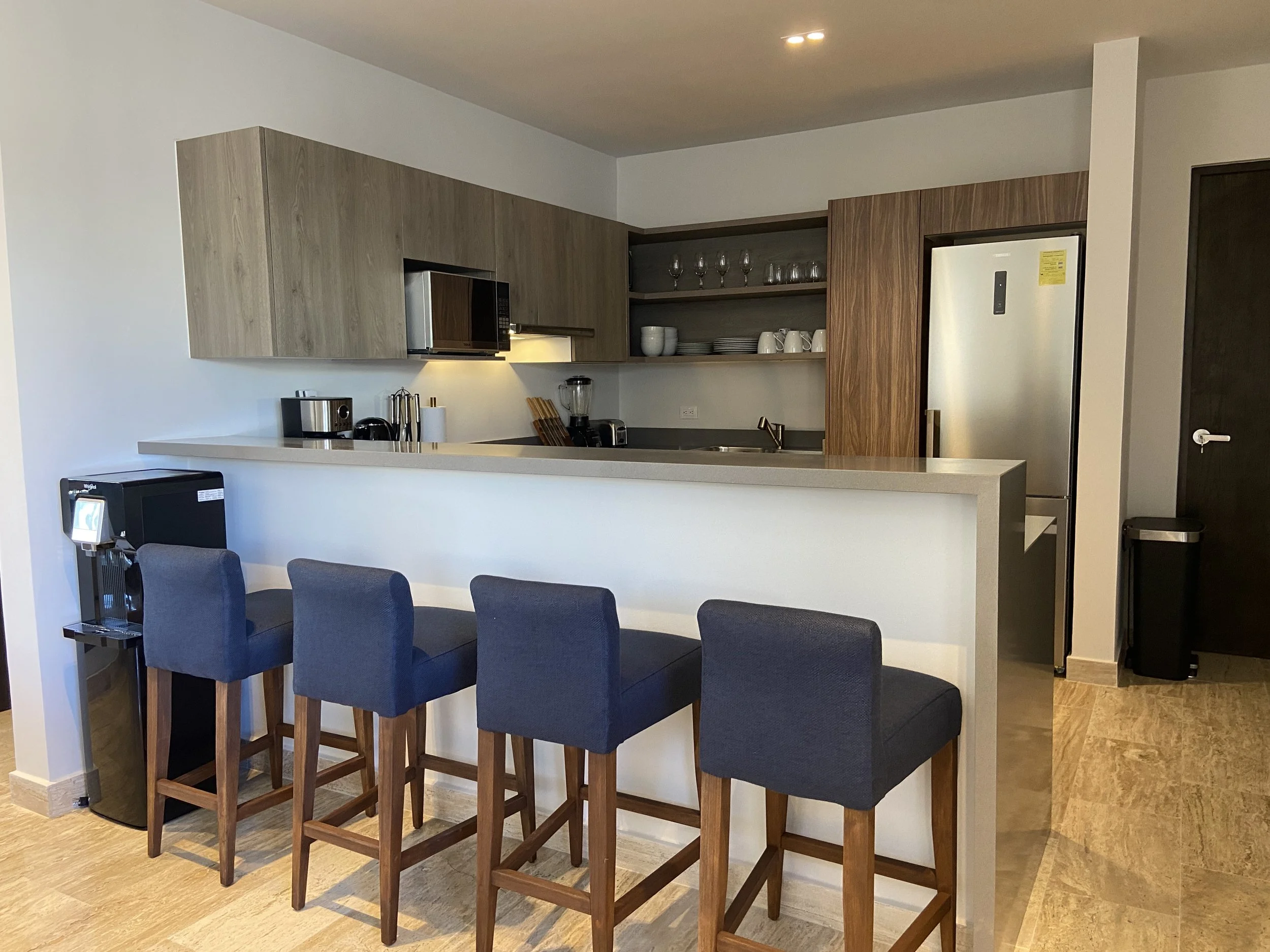Cocina moderna con encimera blanca, electrodomésticos de acero inoxidable, estantes con platos y vasos, y barra con cuatro sillas tapizadas en azul.