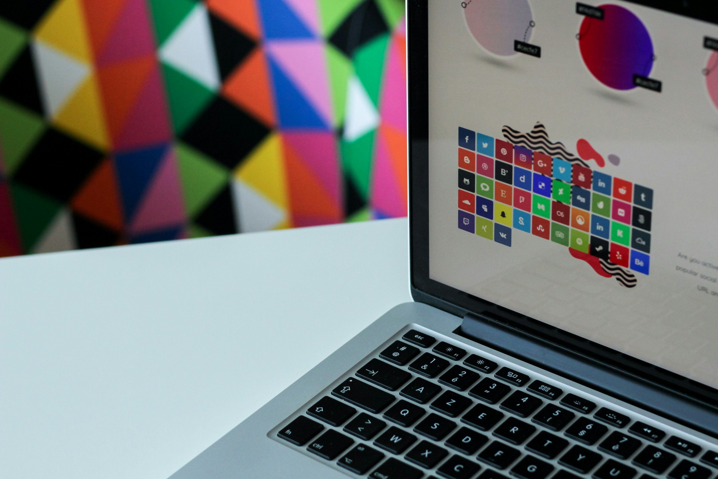 Close-up of a laptop with a colorful social media icons page displayed on the screen. Part of the keyboard is visible at the bottom, and a colorful, geometric patterned object is blurred in the background.