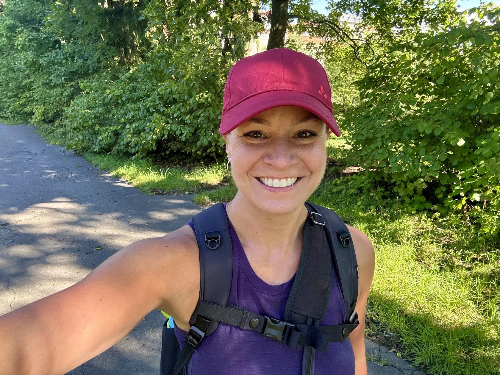 A woman smiling outdoors on a sunny day, wearing a maroon cap, purple athletic shirt, and a black backpack, with green trees and bushes in the background.