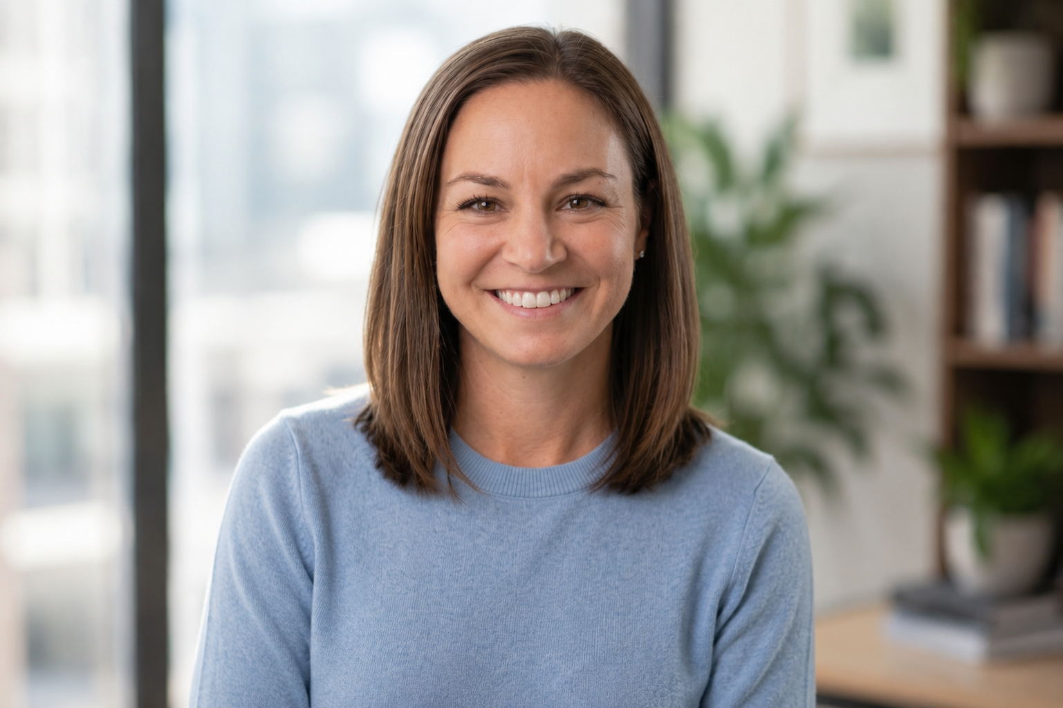 A woman with brown hair, smiling, wearing a blue shirt and navy jacket, standing indoors with blurred plants and windows in the background.