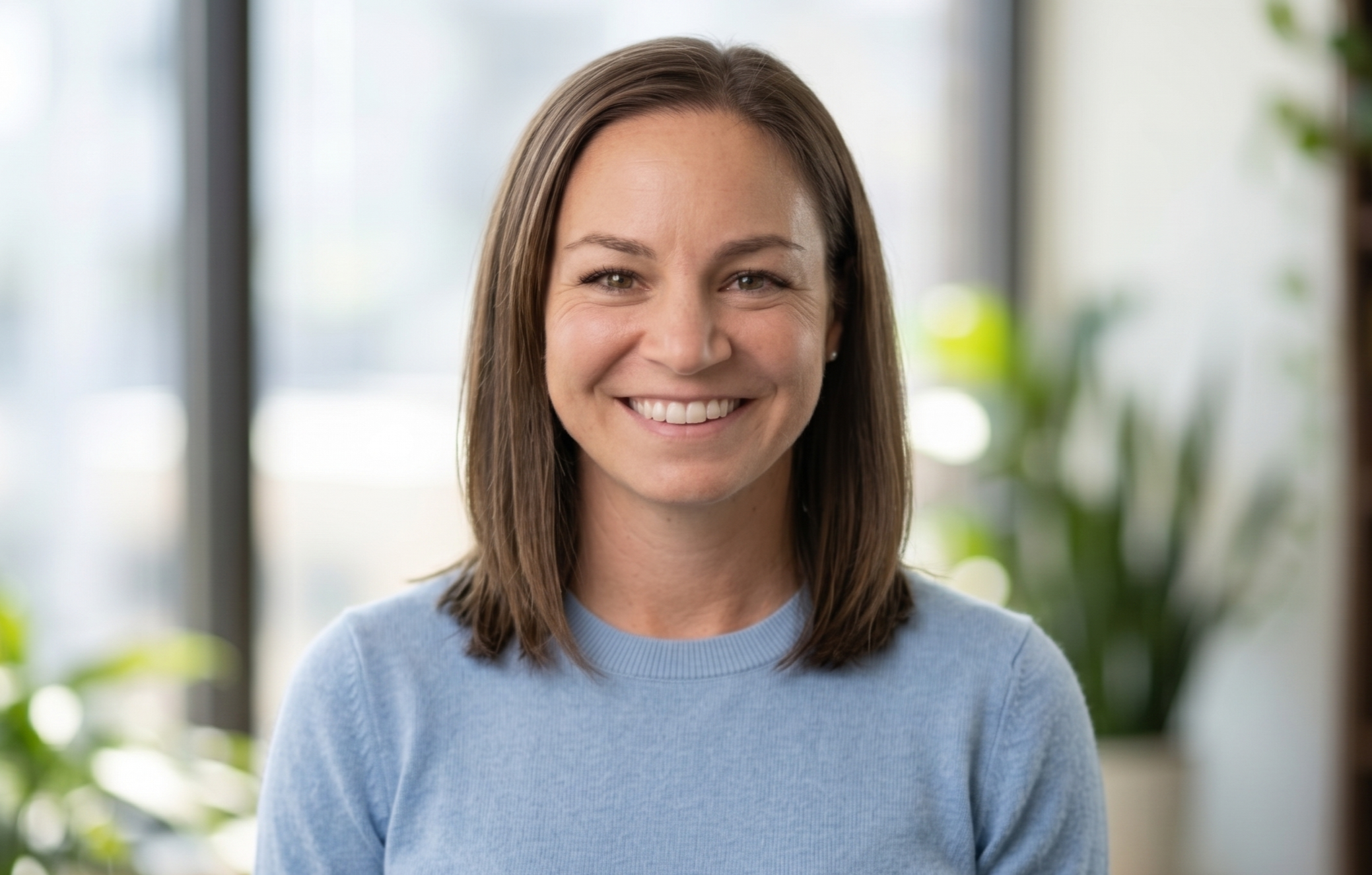 A woman with shoulder-length brown hair wearing a light blue sweater smiling indoors with natural light and blurred background.