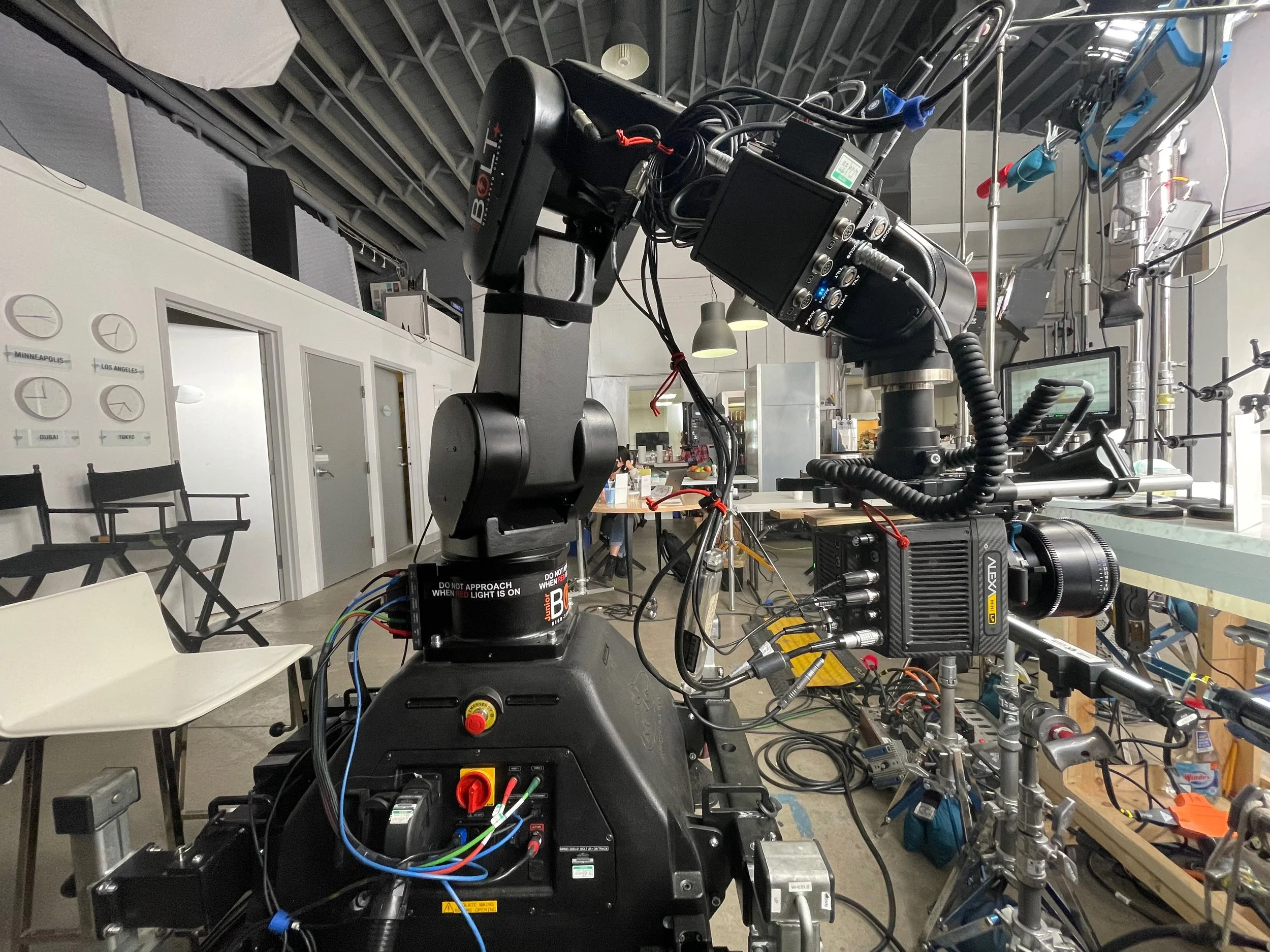 A large robotic arm with multiple circuits and cables connected, situated in a workspace with chairs, tables, and industrial equipment in the background.