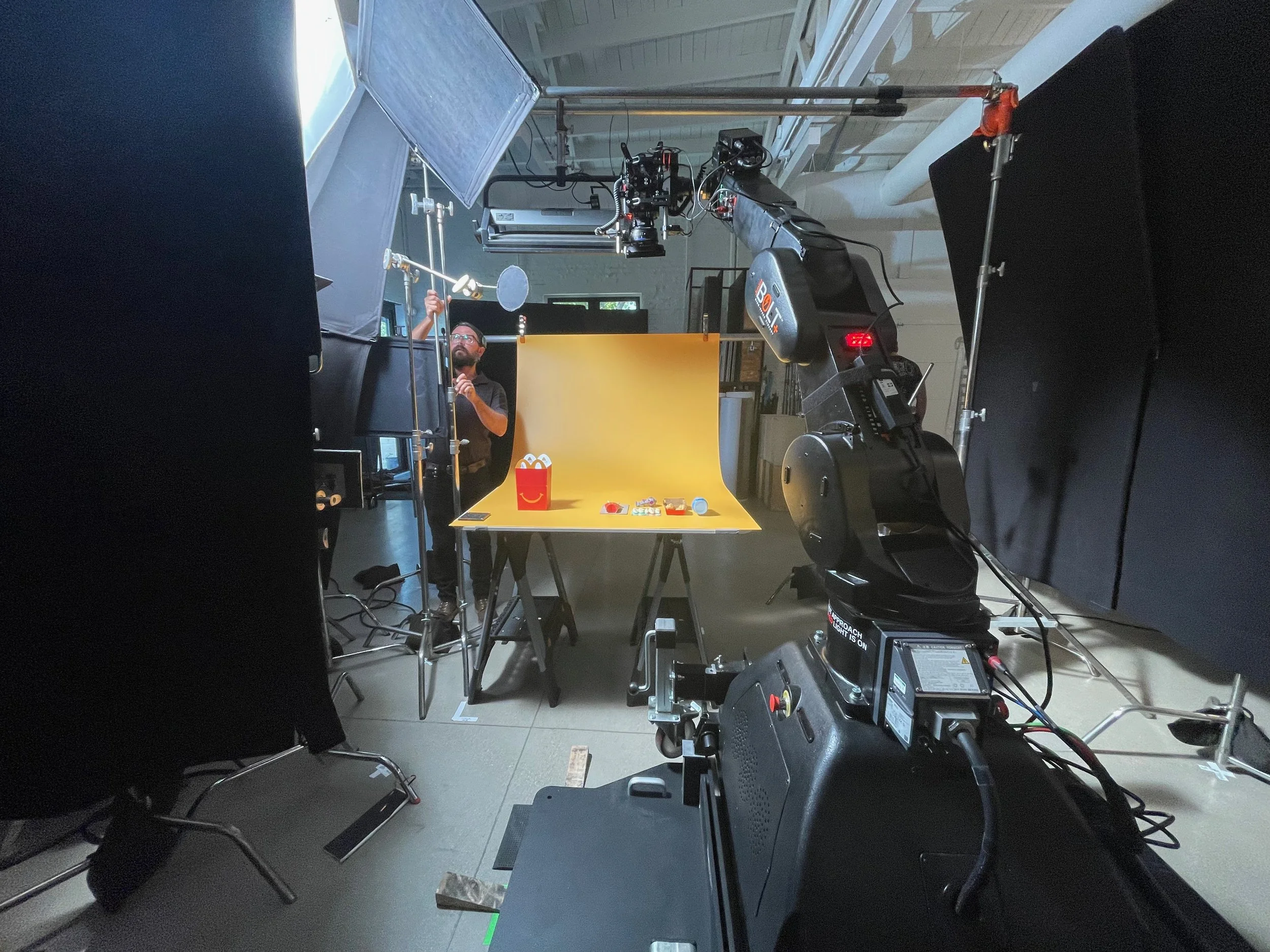 Studio setup with a man adjusting equipment in front of a yellow backdrop, surrounded by cameras, lights, and technical gear.