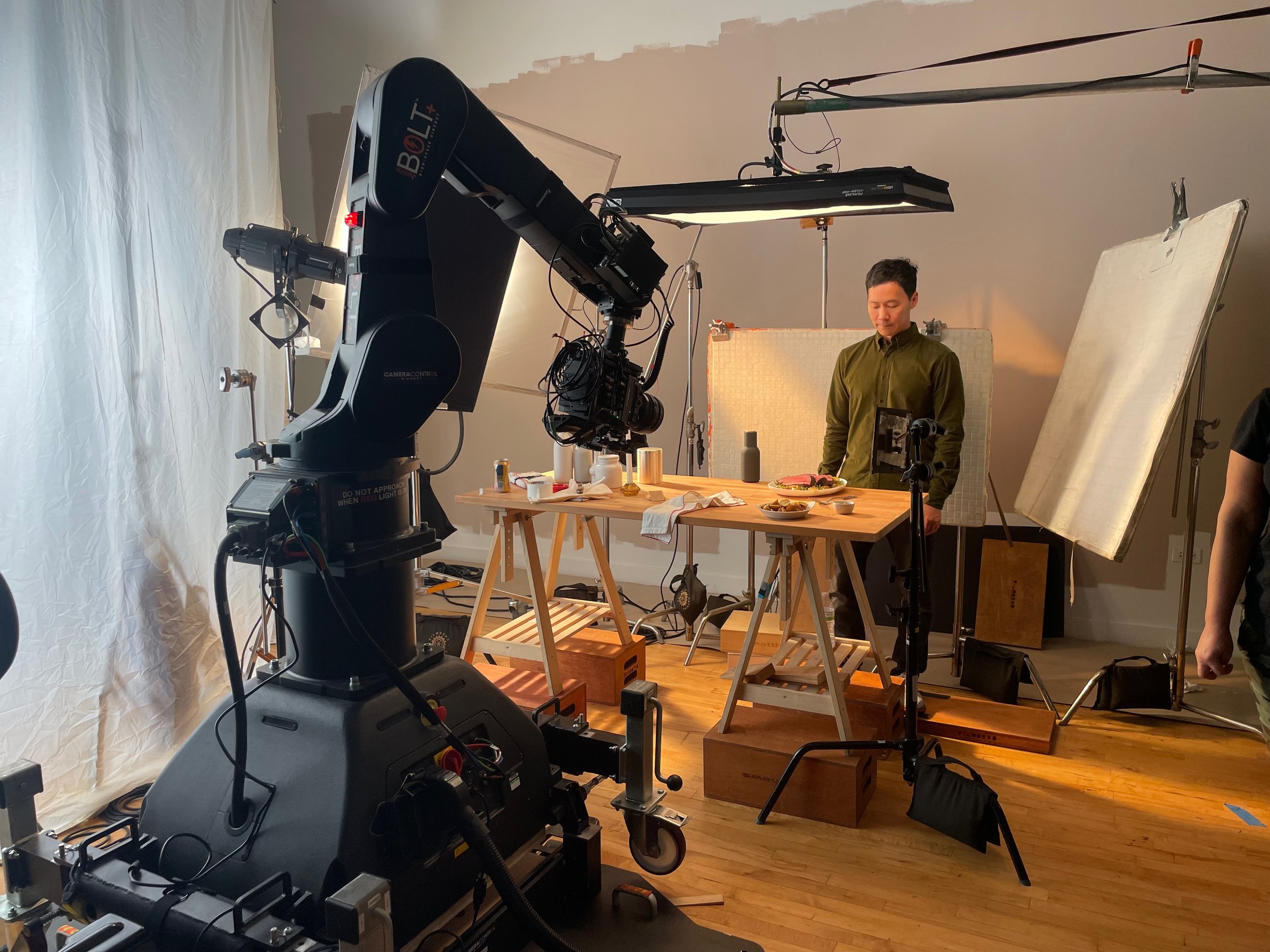 A film or photography set with a large professional camera and lighting equipment, a person standing behind a table with food, and various props and tools scattered around.