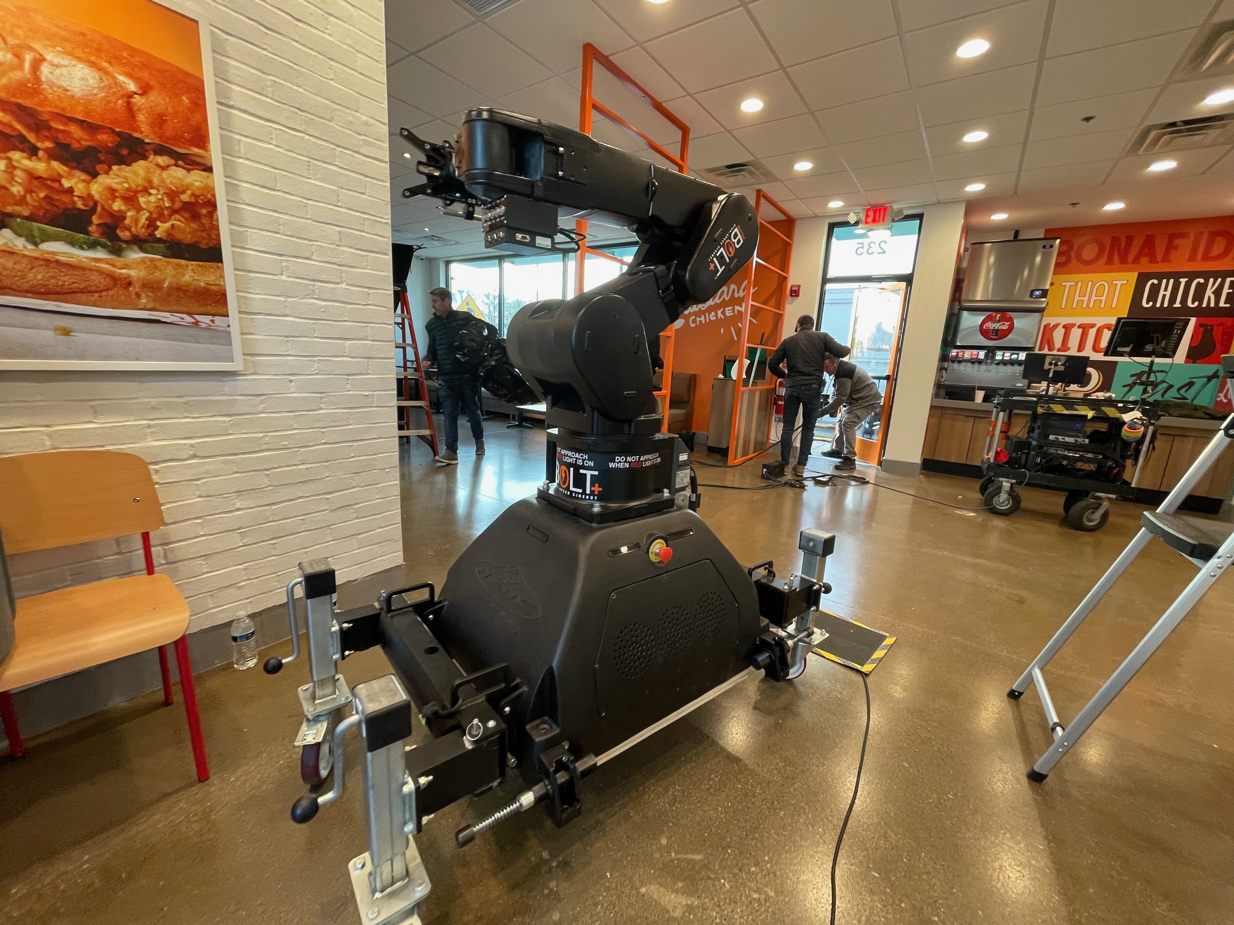 A robot arm in a fast-food restaurant, with a sign on it that says 'BOLT JR'. In the background, there are people and a beverage dispenser, with a colorful wall and window letting in natural light.
