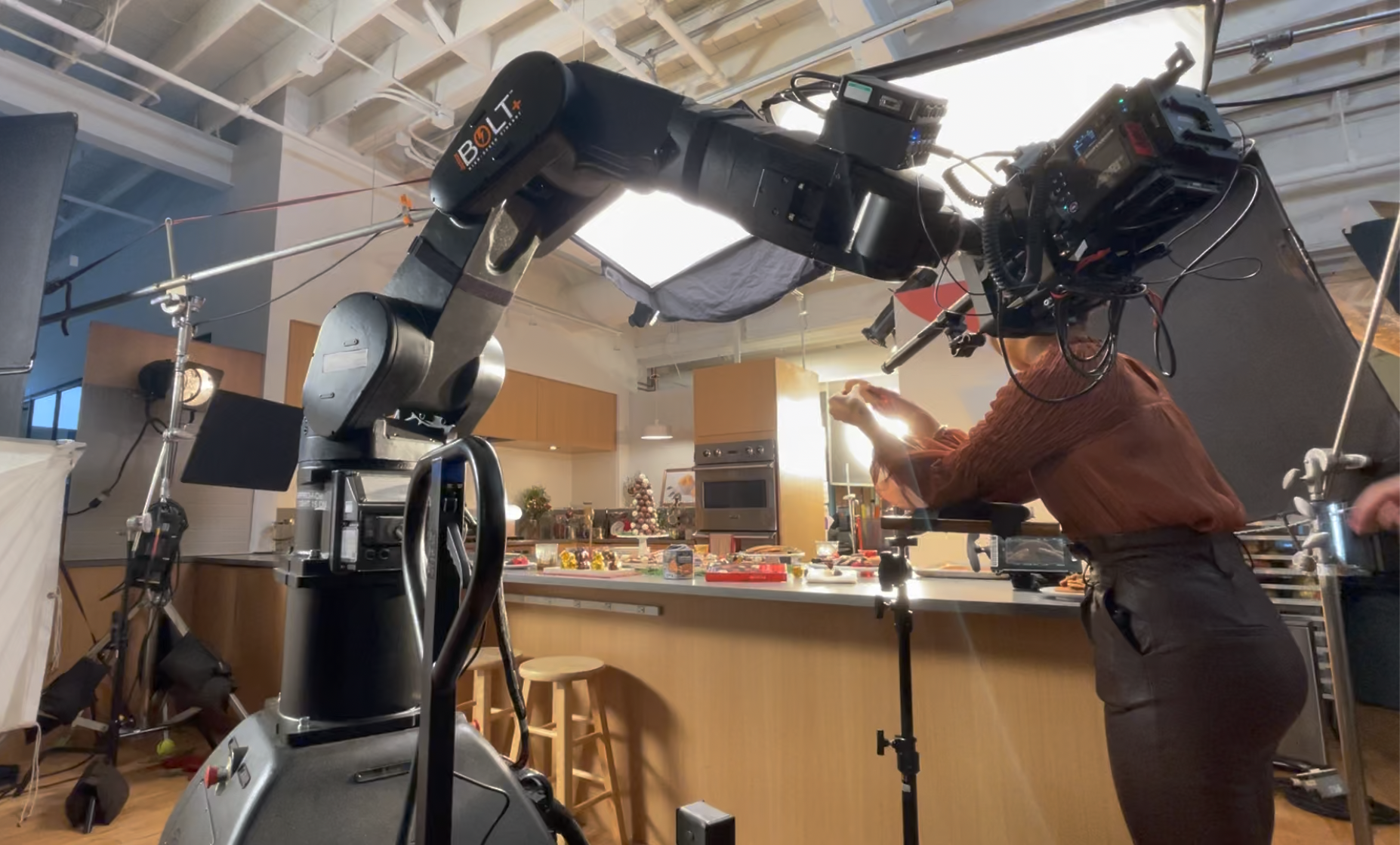 A person in a brown shirt and dark pants stands in a kitchen, adjusting lighting equipment while a robotic arm or camera setup is positioned nearby. The kitchen has wooden cabinets, a stainless steel oven, and holiday decorations on the counter, sugg