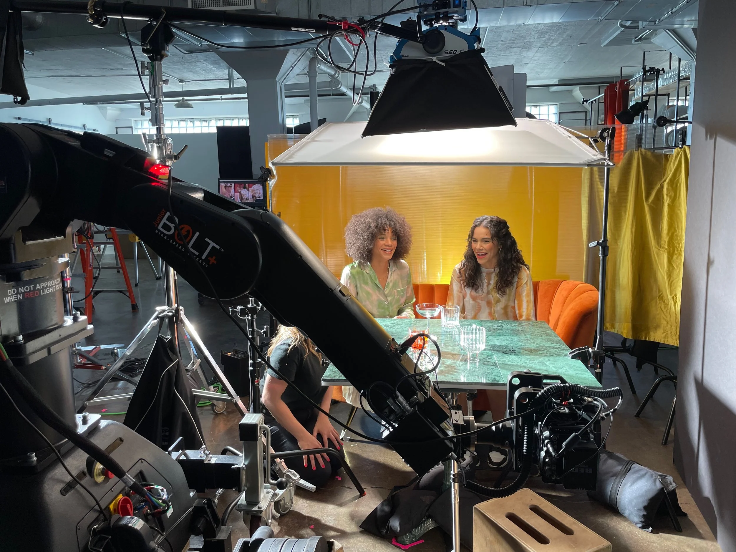 Two women sitting at a table during a photoshoot, with professional lighting and filming equipment visible around them in a modern studio setting.