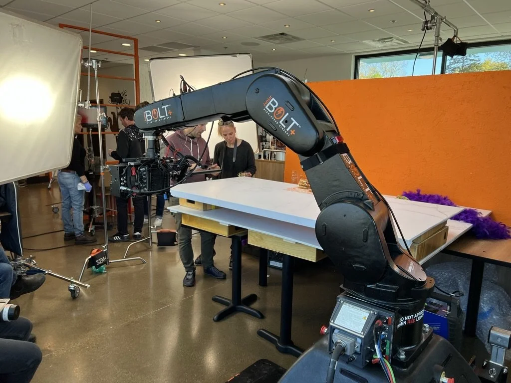 A large black robotic arm labeled 'Bolt' is in a studio, holding a white flat surface on a wooden table, with people observing and working around it. The studio has equipment, lights, and a bright orange backdrop.