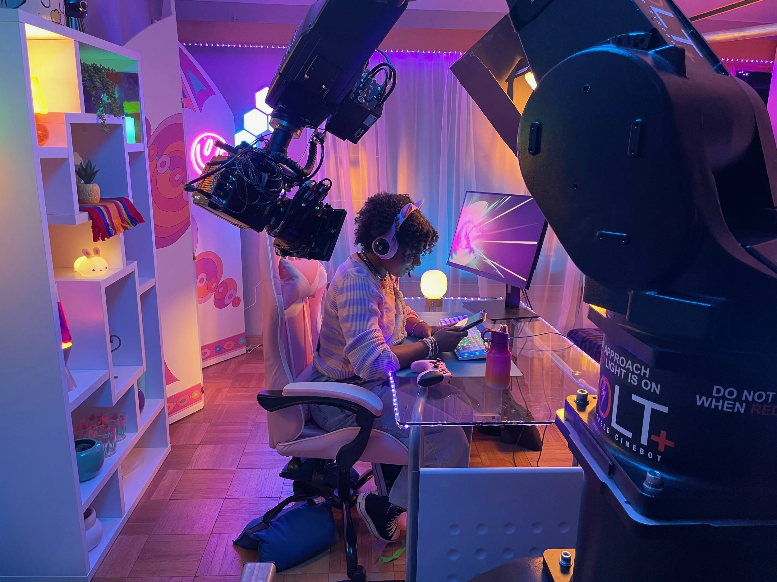 A woman with curly hair wearing headphones and a light-colored striped shirt, seated at a desk with a digital music controller. A large camera on a crane is positioned in front of her, capturing her on video. The background has colorful neon lights, 