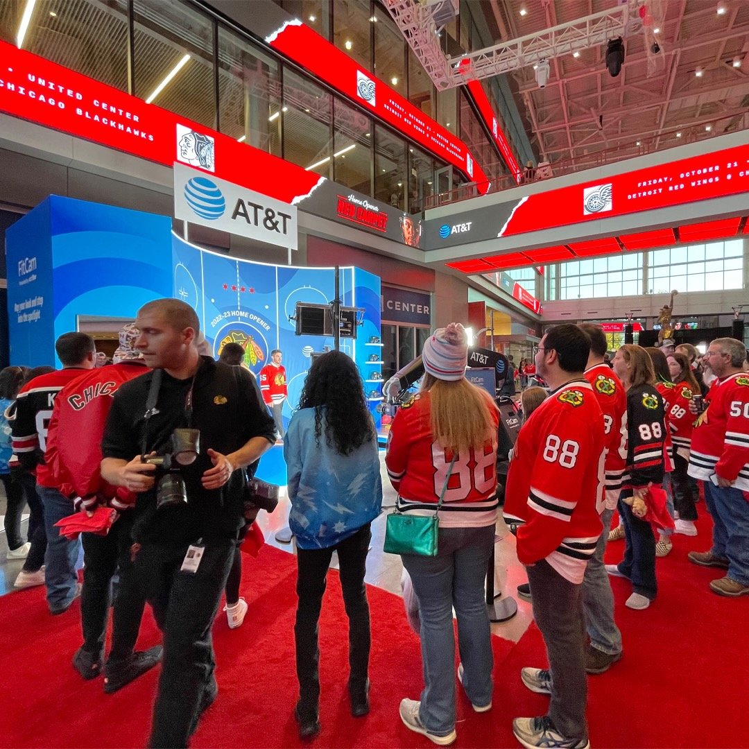 A crowd of people wearing Chicago Blackhawks hockey jerseys inside the United Center in Chicago, Illinois, during an event. The venue features red electronic signs, a blue AT&T display, and hockey-related decorations.