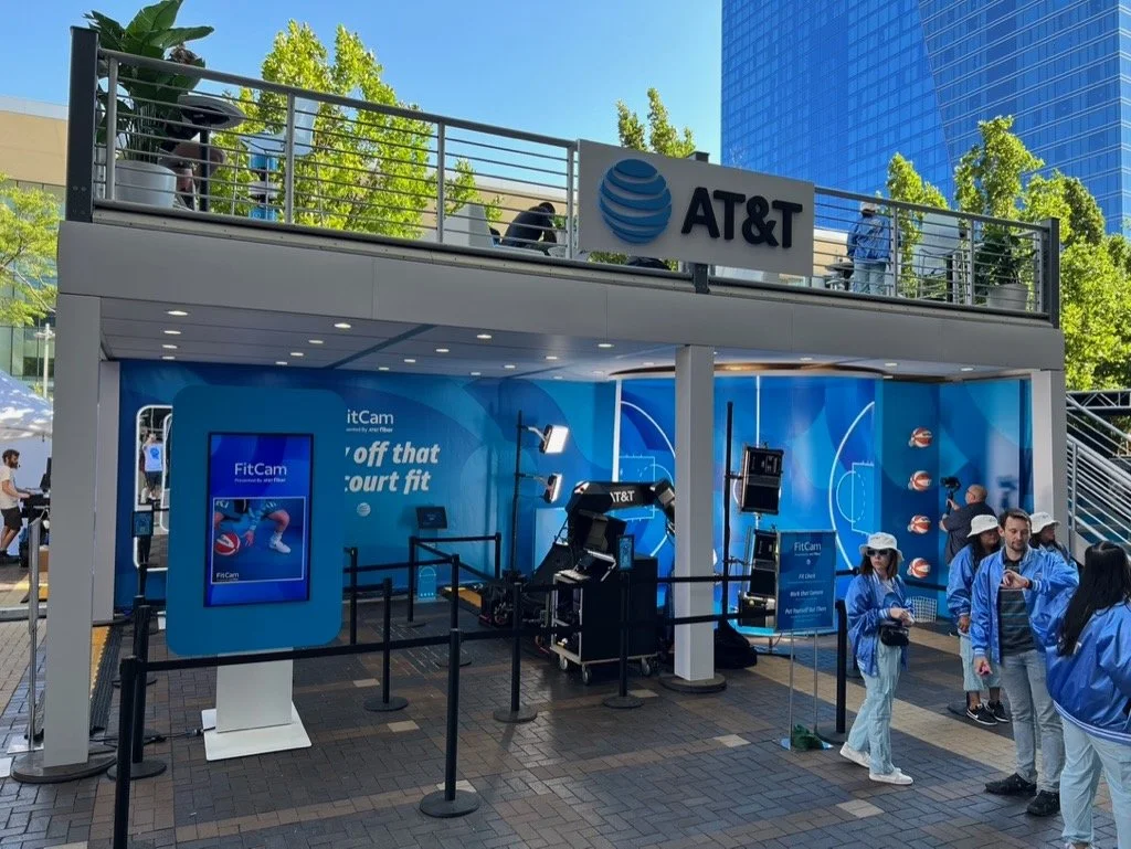 AT&T promotional booth featuring fitness technologies like FitCam and itCam at an outdoor event. The booth has a large digital screen displaying FitCam, and people wearing blue AT&T jackets are gathered around. There are trees and tall buildings in t