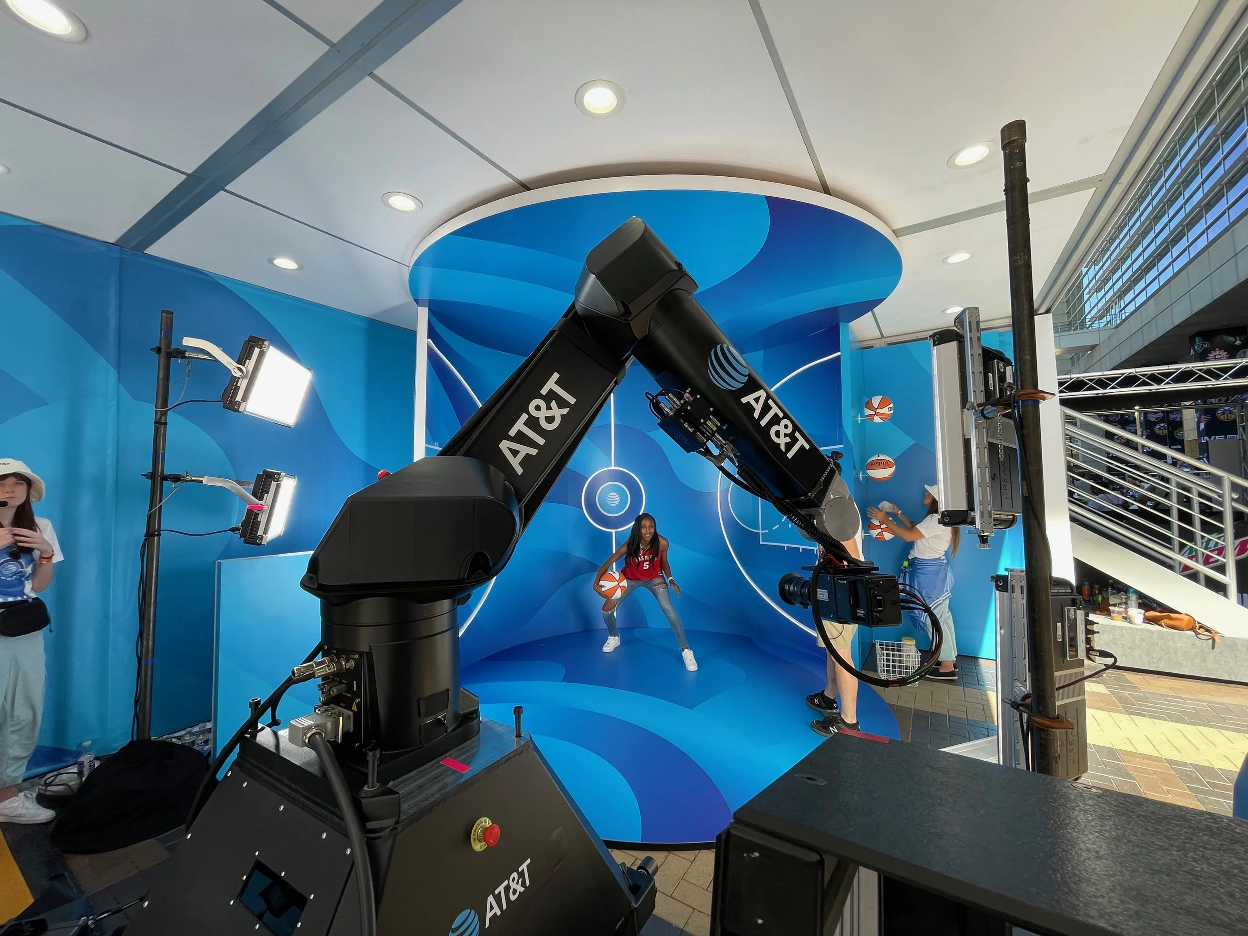 A camera rig and lighting equipment set up in front of a blue backdrop at an indoor event. Two women are visible, one smiling at the camera and the other holding a basketball. The scene appears to be a photo or video shoot area.