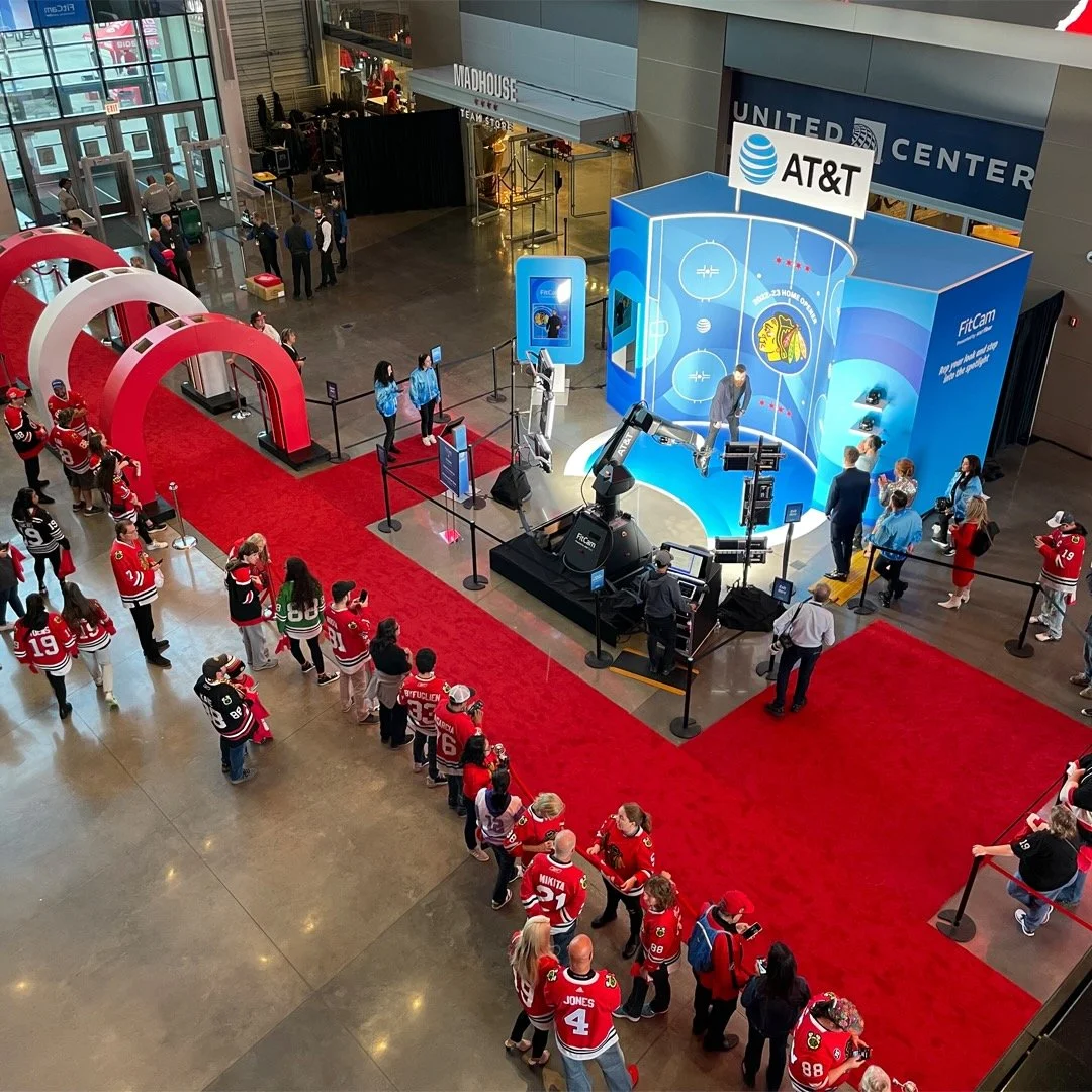 People in hockey jerseys queue to participate in AT&T hockey-themed virtual experience in a shopping mall, with a red carpet, hockey-related displays, and a robotic arm.