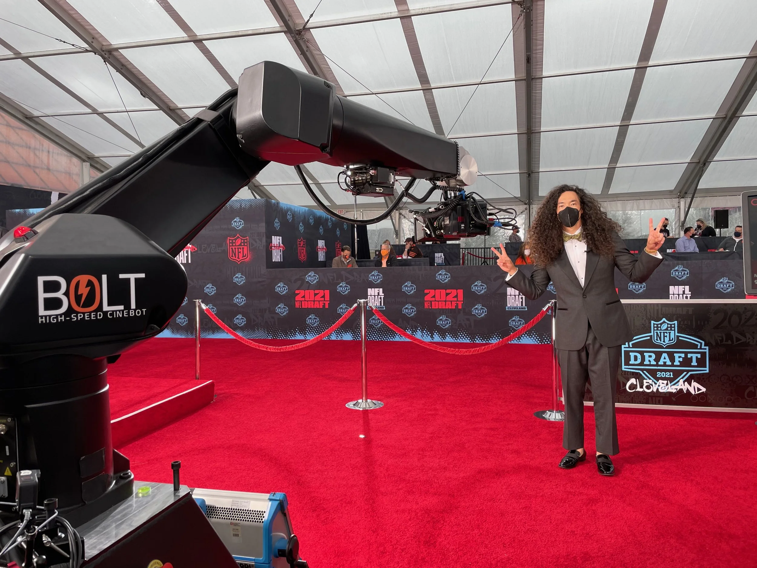 A person in a tuxedo and face mask smiling and making peace signs at a 2021 NFL Draft event, with a large camera and camera operator in the foreground and event branding in the background.