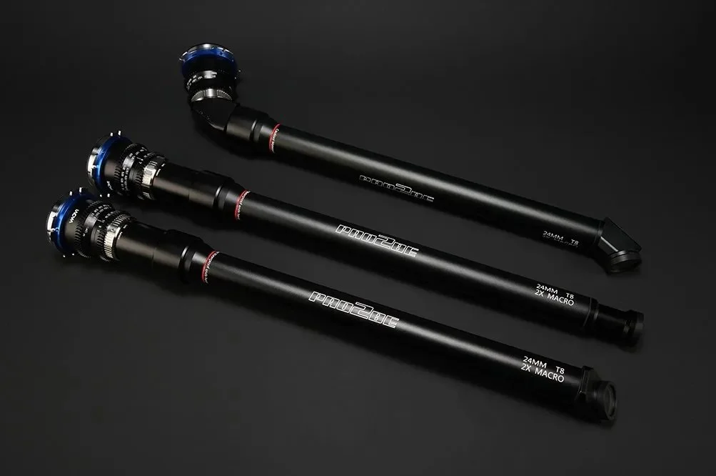 Three black camera lens rods with blue crown mounts, labeled 'PRO2BE', '24MM T8 2X MACRO', on a dark background. Laowa