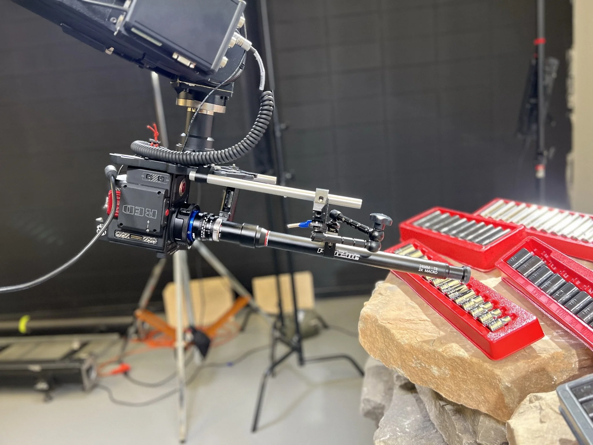 Close-up of a professional camera mounted on a tripod, aimed at a workbench with a red toolbox containing camera lenses and equipment, in a studio setting.