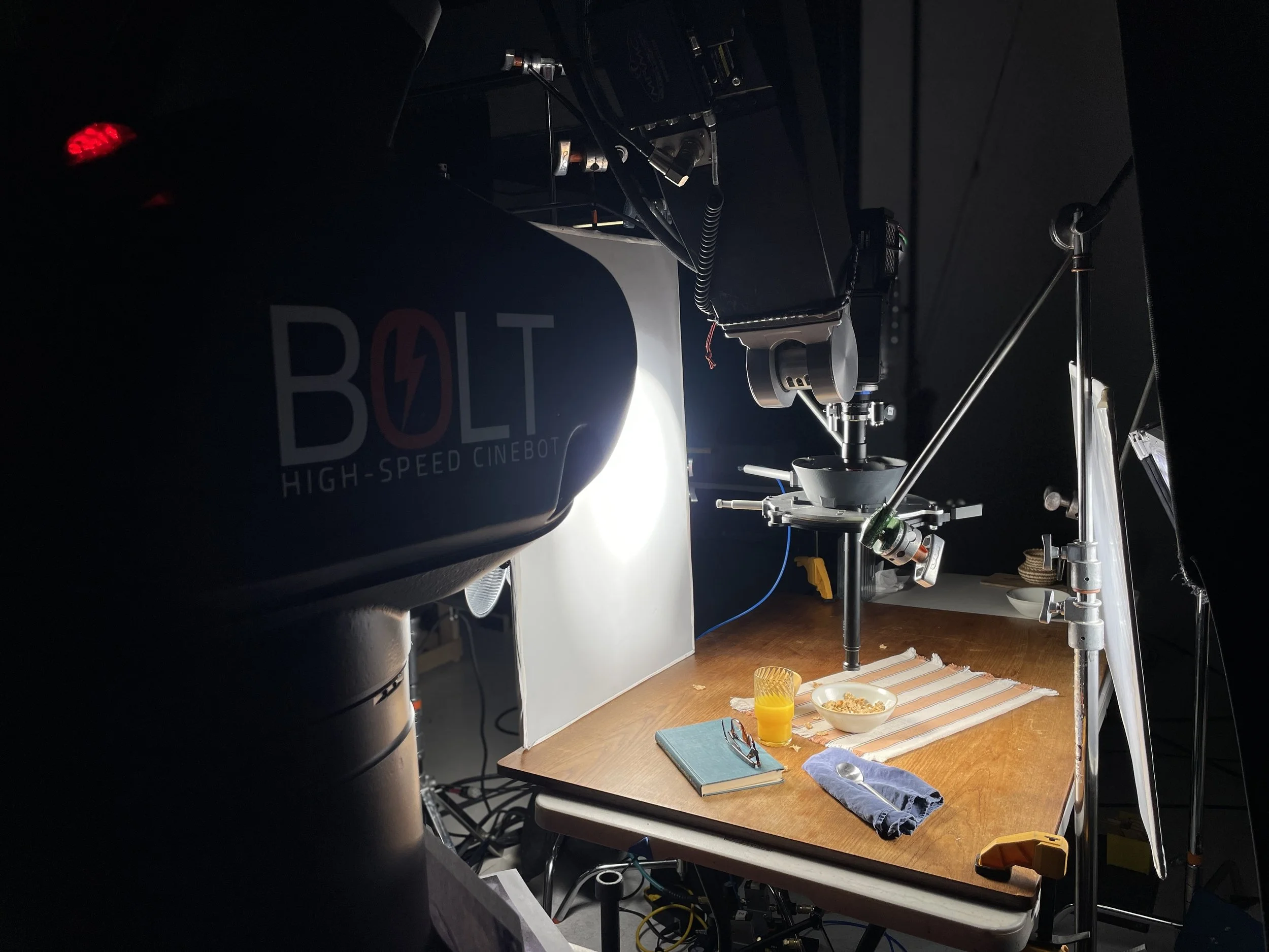 A photography studio setup with a table that has a bowl of cereal, a glass of orange juice, a notebook, glasses, a cloth, and a spoon. The background shows a black wall with a logo that reads 'BOLT High-Speed Cinebot' and some studio equipment and li