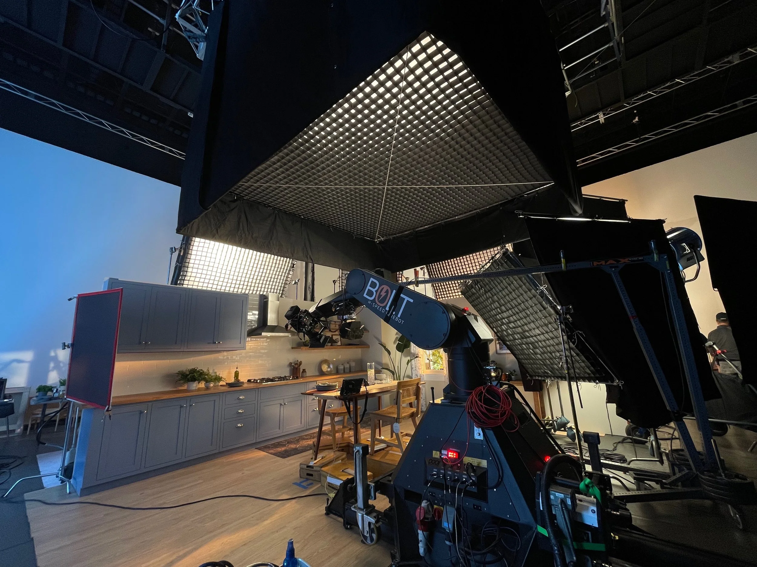 A film or television set with lighting and equipment, set in a home interior featuring kitchen cabinets and a dining table.