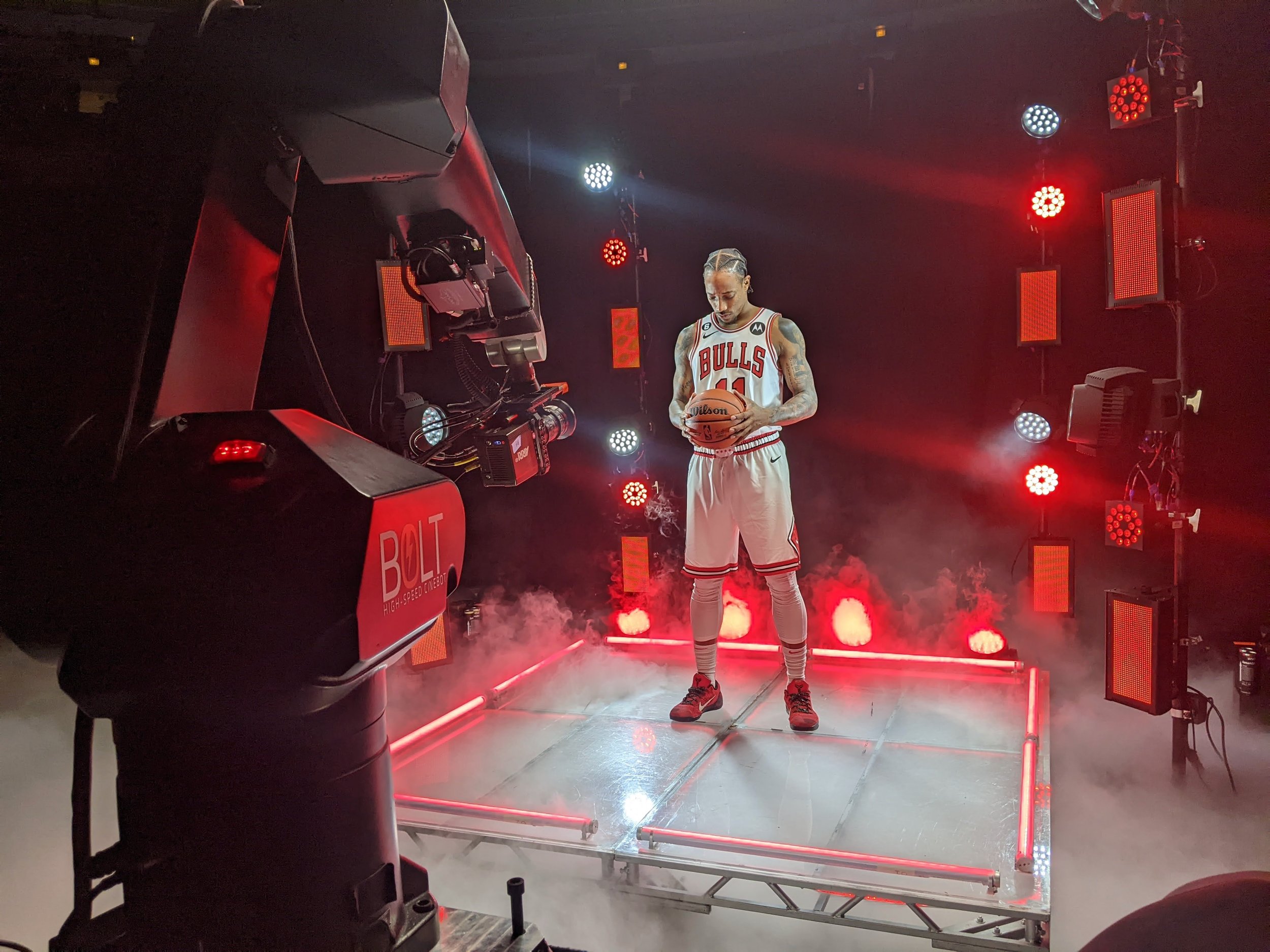 A person wearing a Chicago Bulls basketball uniform holding a basketball standing on a platform surrounded by red and white stage lights and smoke effects.